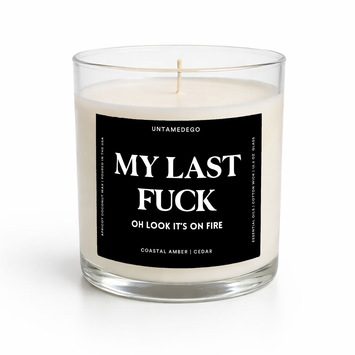 A white soy wax candle in a clear glass jar with a black square label that reads 'MY LAST FUCK OH LOOK IT'S ON FIRE' and smaller text 'UNTAMEDEGO' and 'COASTAL AMBER | CEDAR'.