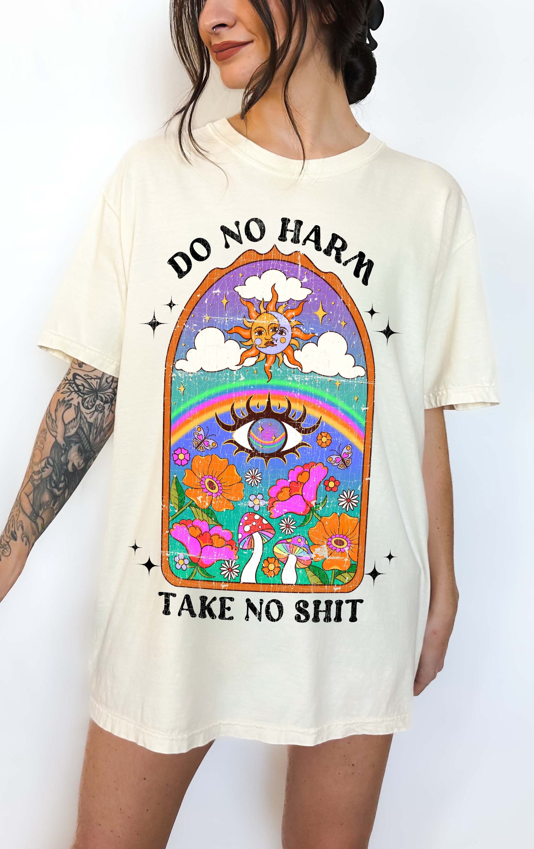 Person wearing Ivory graphic t-shirt featuring a colorful illustrated scene with sun, rainbow, flowers, mushrooms, and the text Do No Harm Take No Shit, photographed against a light background