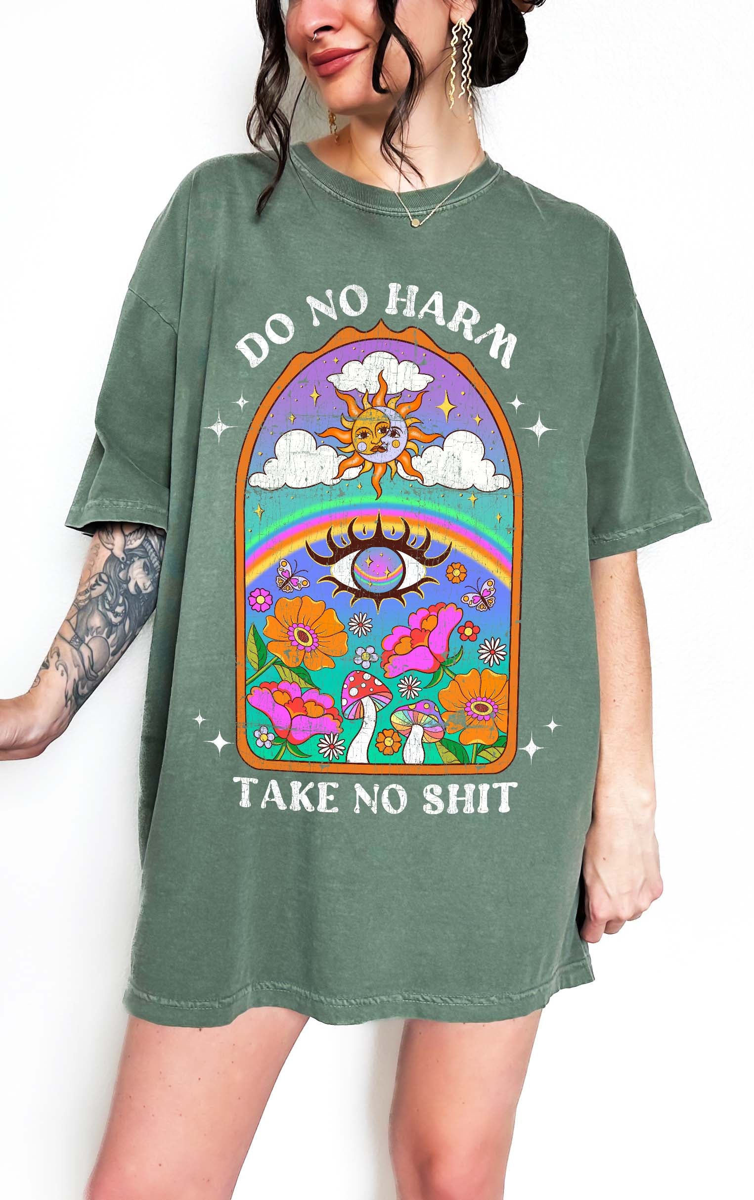 Person wearing a green graphic t-shirt featuring a colorful illustrated scene with sun, rainbow, flowers, mushrooms, and the text Do No Harm Take No Shit, photographed against a light background