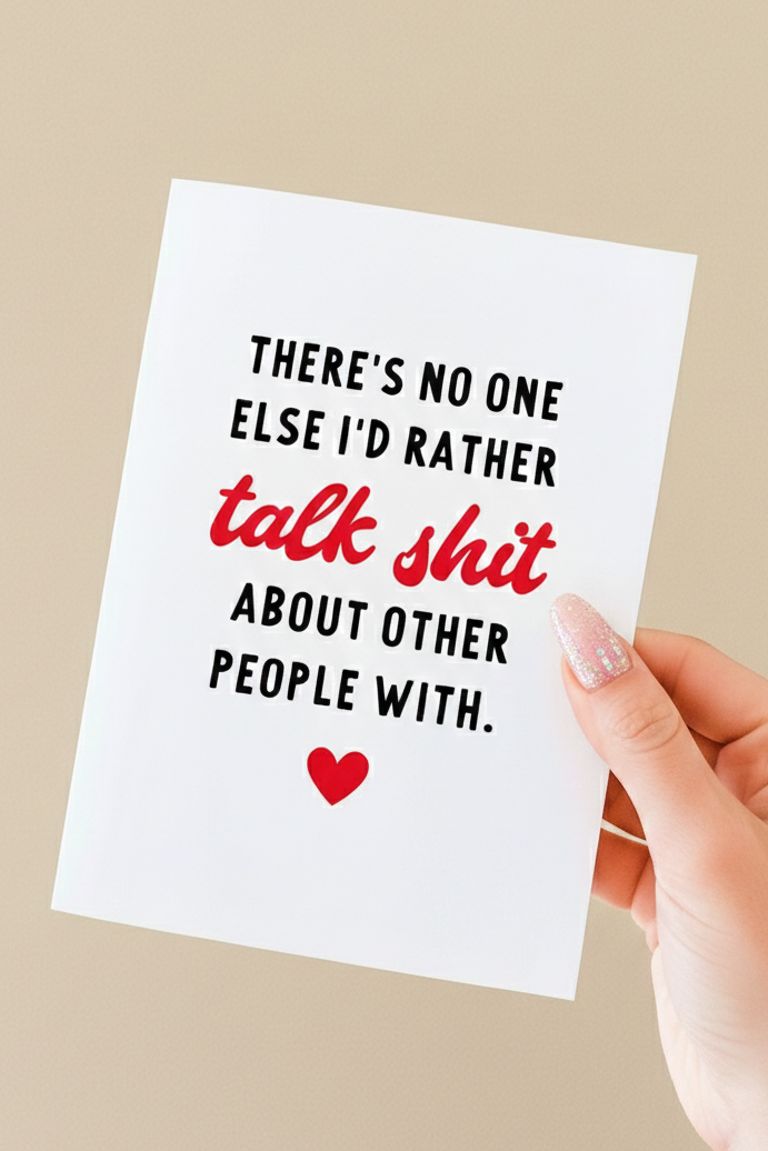 Greeting card held in a hand against a neutral background. The card has a white background with bold black text that reads, “There’s no one else I’d rather,” followed by the phrase “talk shit” in red script-style lettering, and then black text reading, “about other people with.” A small red heart icon appears centered near the bottom of the card. The card is vertically oriented and blank inside