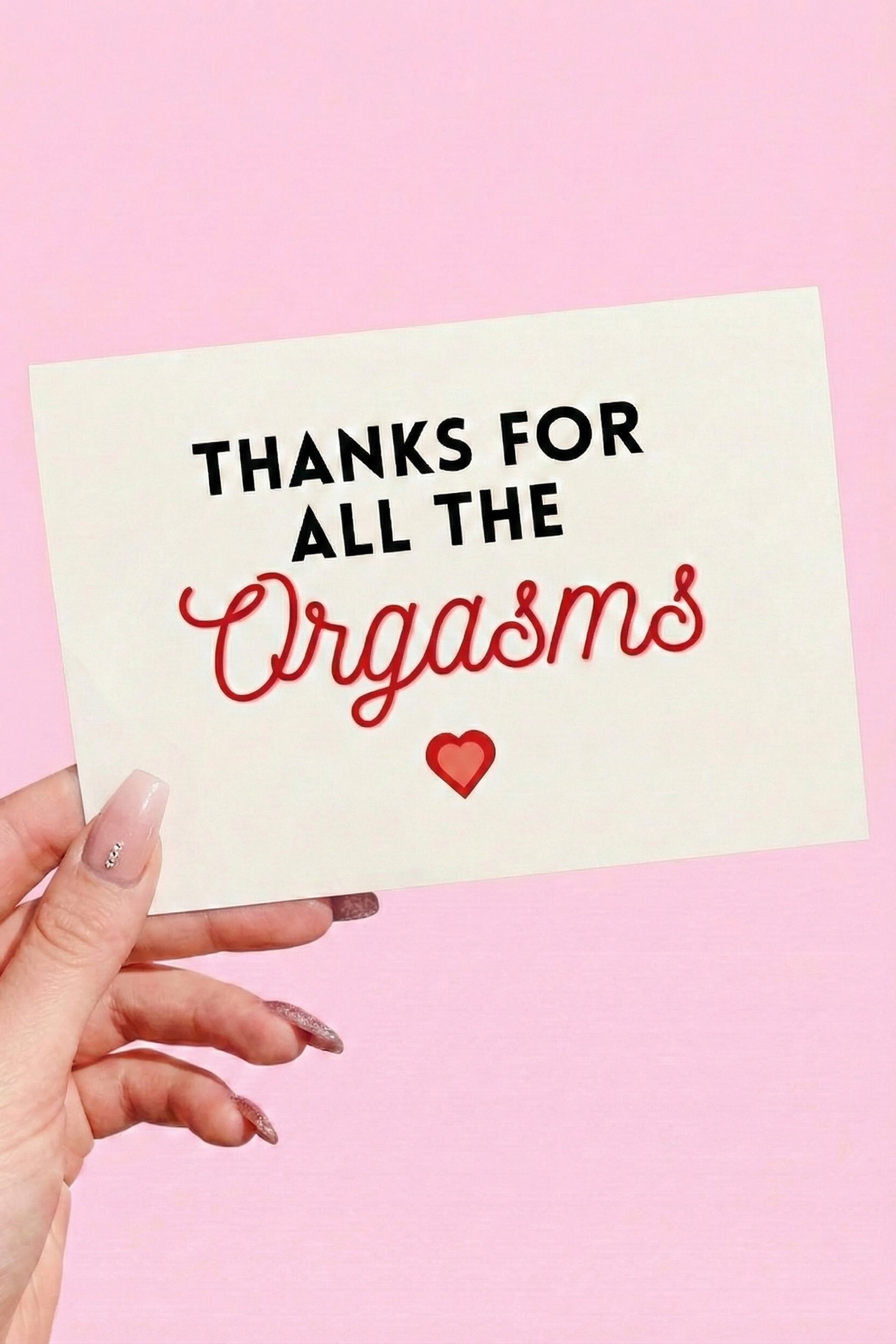 Greeting card with black and red text and a heart graphic on a light pink background, photographed being held by a hand