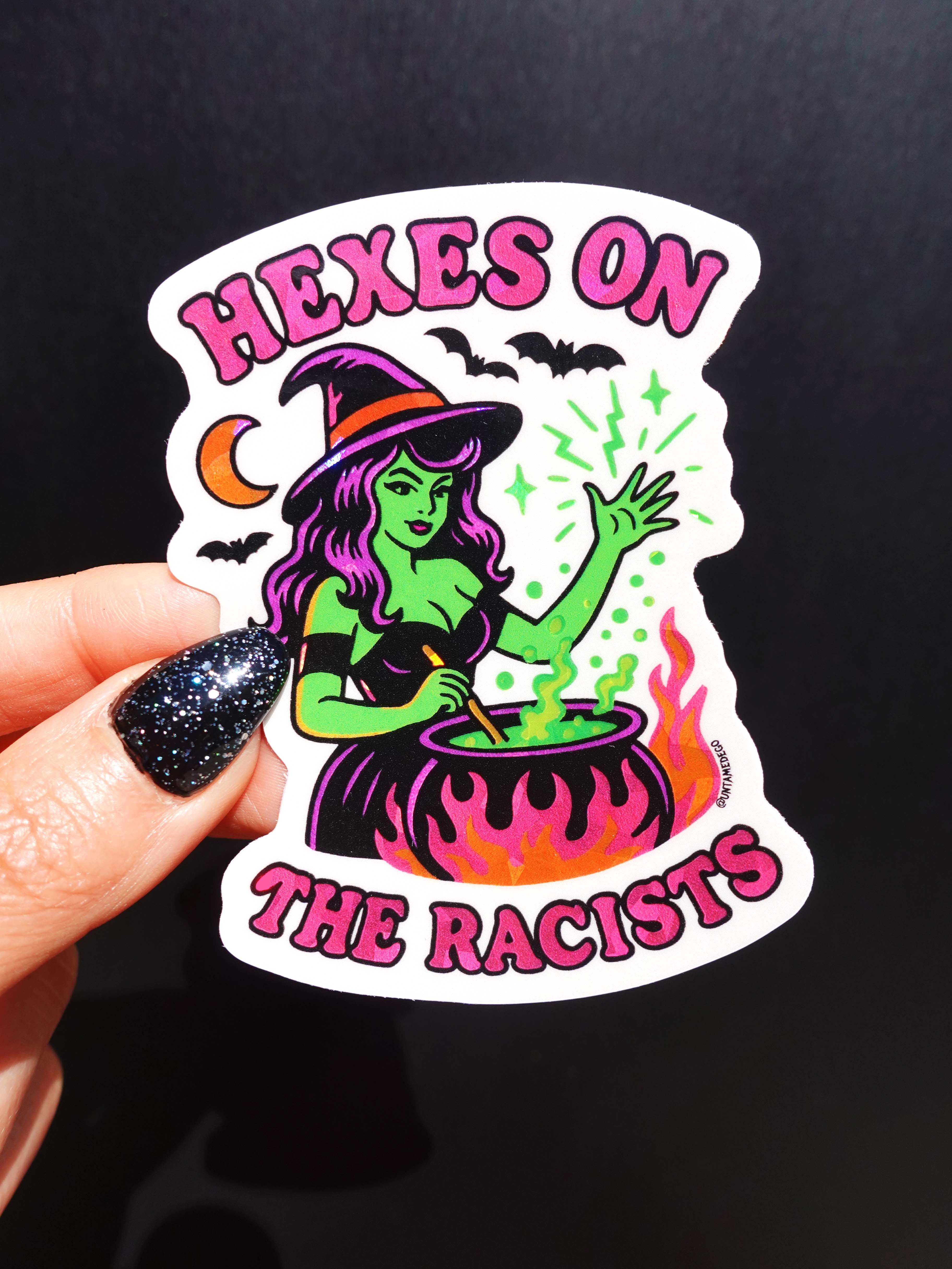 Sticker with a witch design and text 'Hexes on the Racists' held by a hand against a black background.