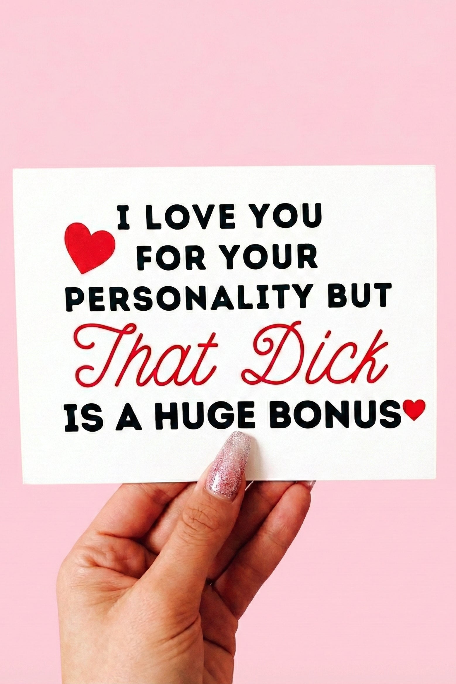 Greeting card displaying a humorous adult phrase in black and red text with heart graphics, photographed being held by a hand against a pink background