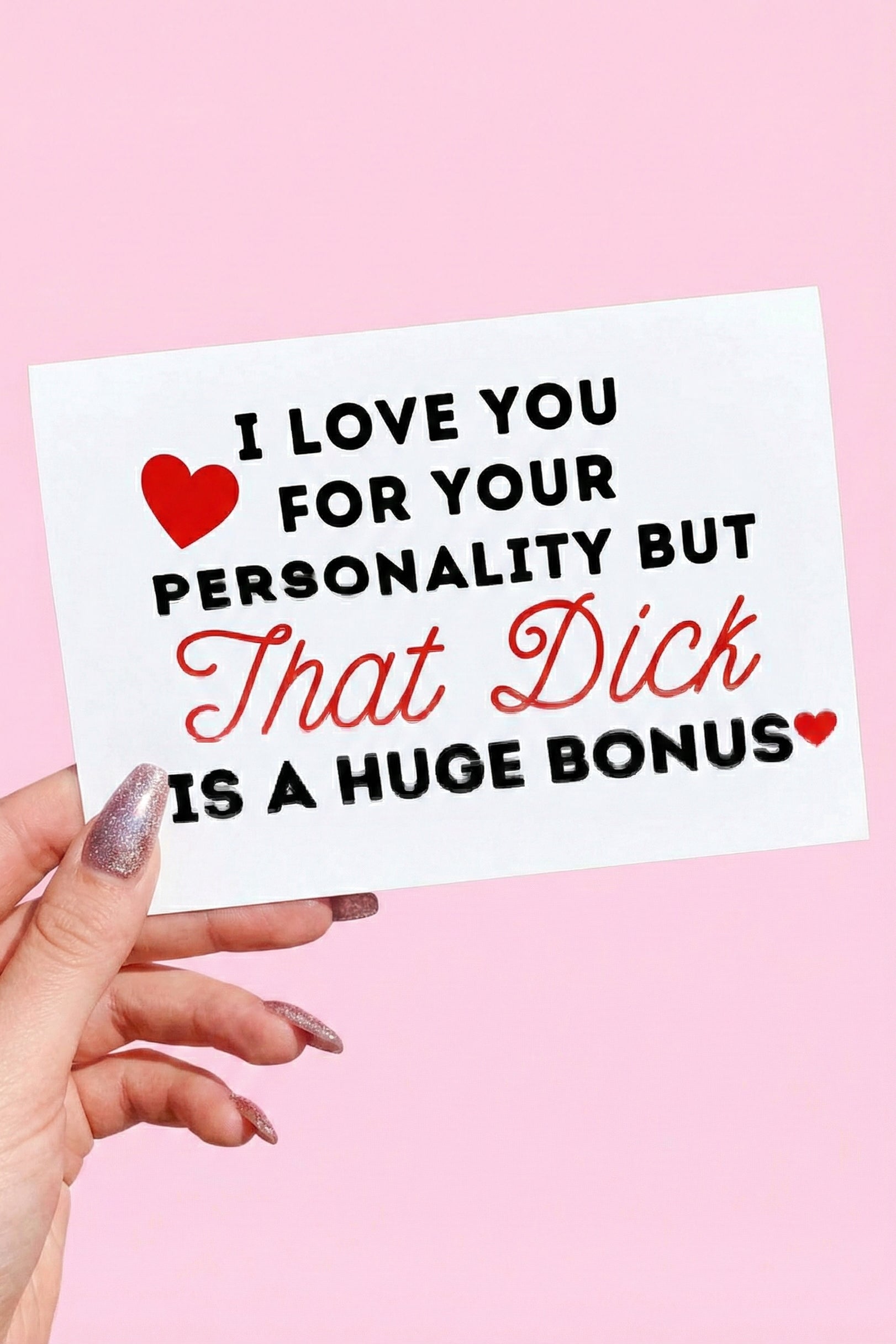 Greeting card displaying a humorous adult phrase in black and red text with heart graphics, photographed being held by a hand against a pink background