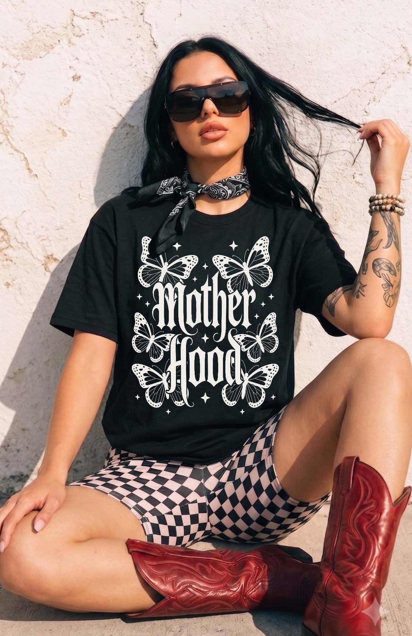 A woman wearing a black graphic t-shirt with 'Mother Hood' and butterflies, black-and-white checkered shorts, and red cowboy boots, sitting against a textured white wall.