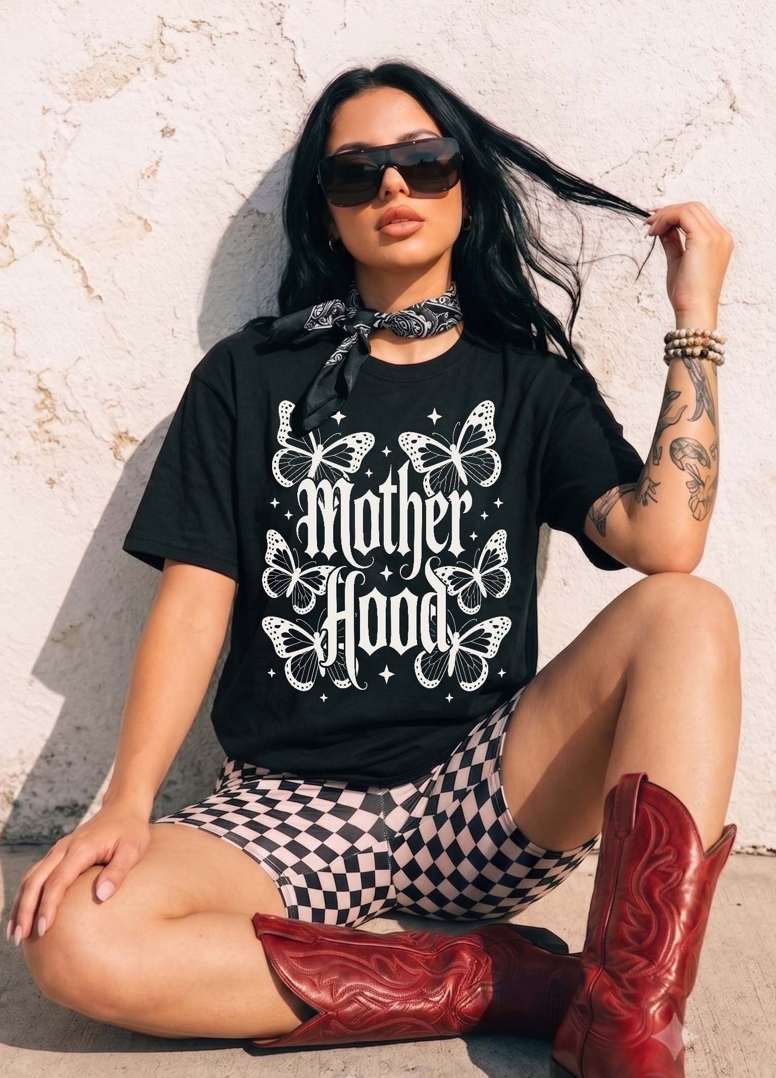 A woman wearing a black graphic t-shirt with 'Mother Hood' and butterflies, black-and-white checkered shorts, and red cowboy boots, sitting against a textured white wall.