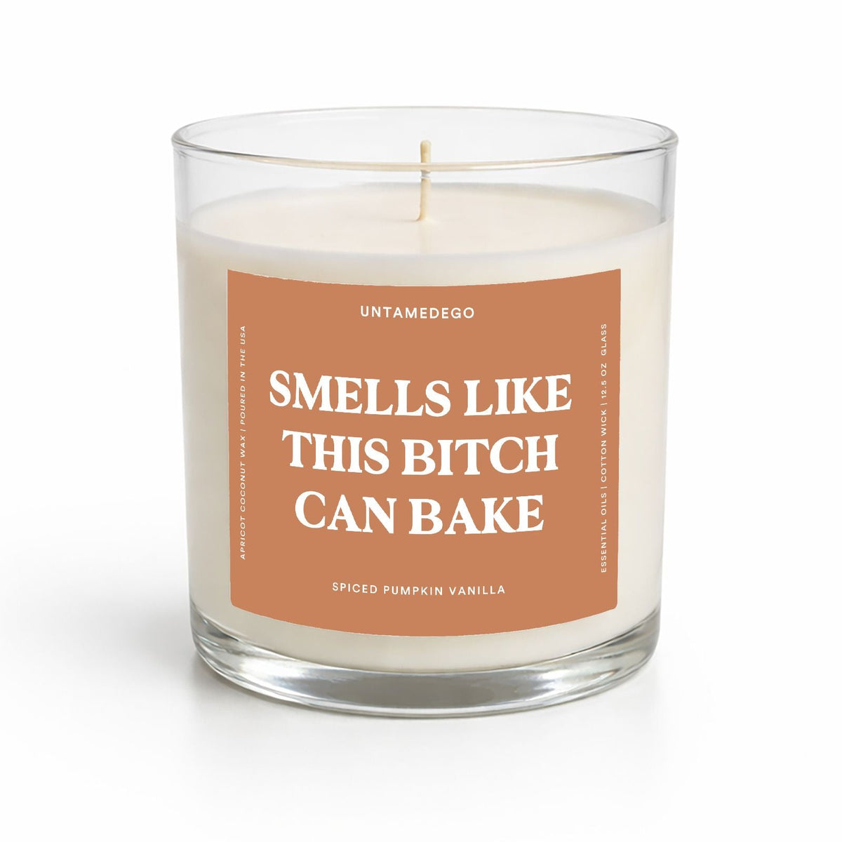 A clear glass jar candle with white wax and a rectangular terracotta label that reads 'SMELLS LIKE THIS BITCH CAN BAKE' in white text. The brand name 'UNTAMEDEGO' is visible at the top of the label, and 'Spiced Pumpkin Vanilla' is written at the bottom.