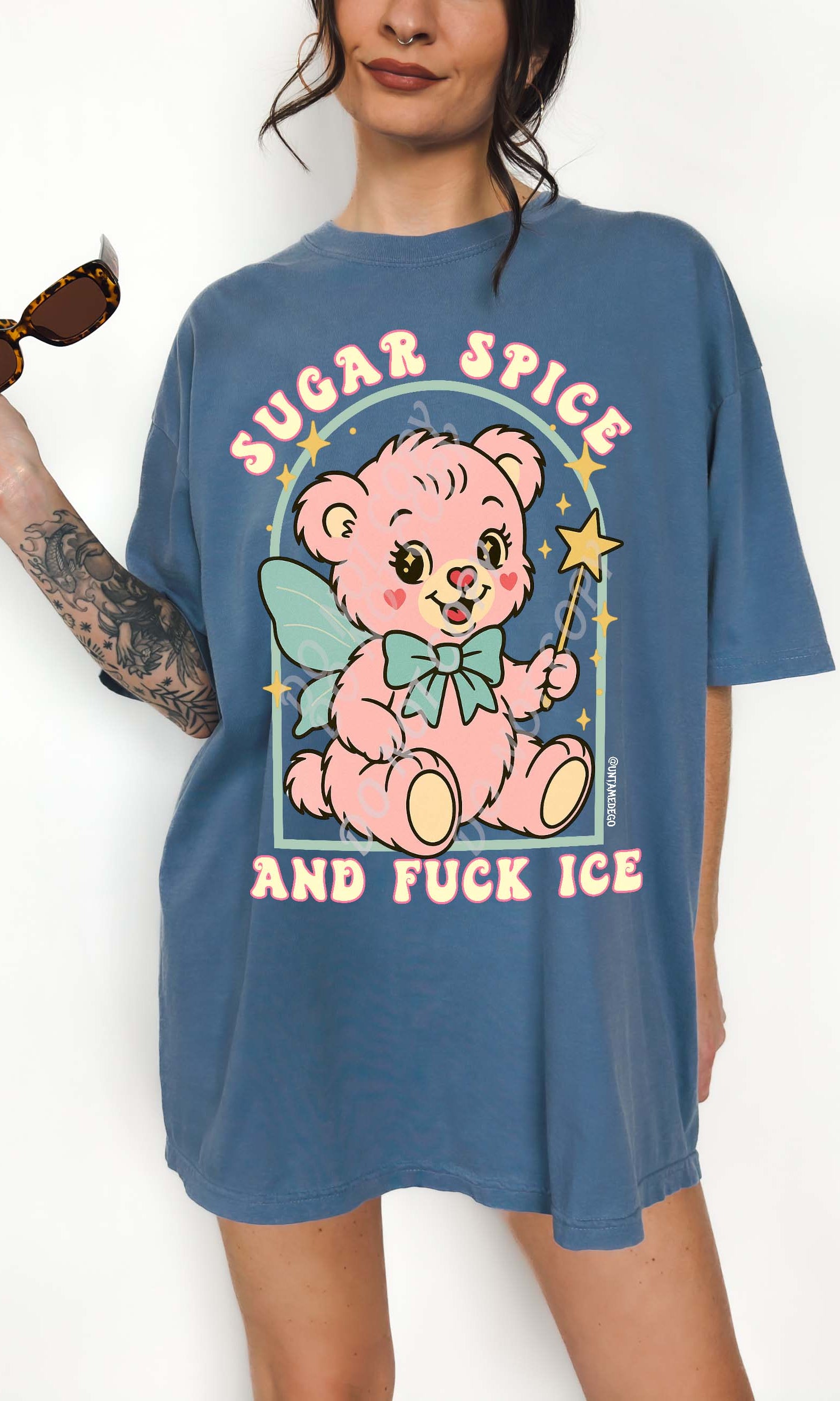Person wearing a blue graphic t-shirt featuring a pastel illustrated bear holding a wand and text that reads Sugar Spice and Fuck ICE, photographed against a white background