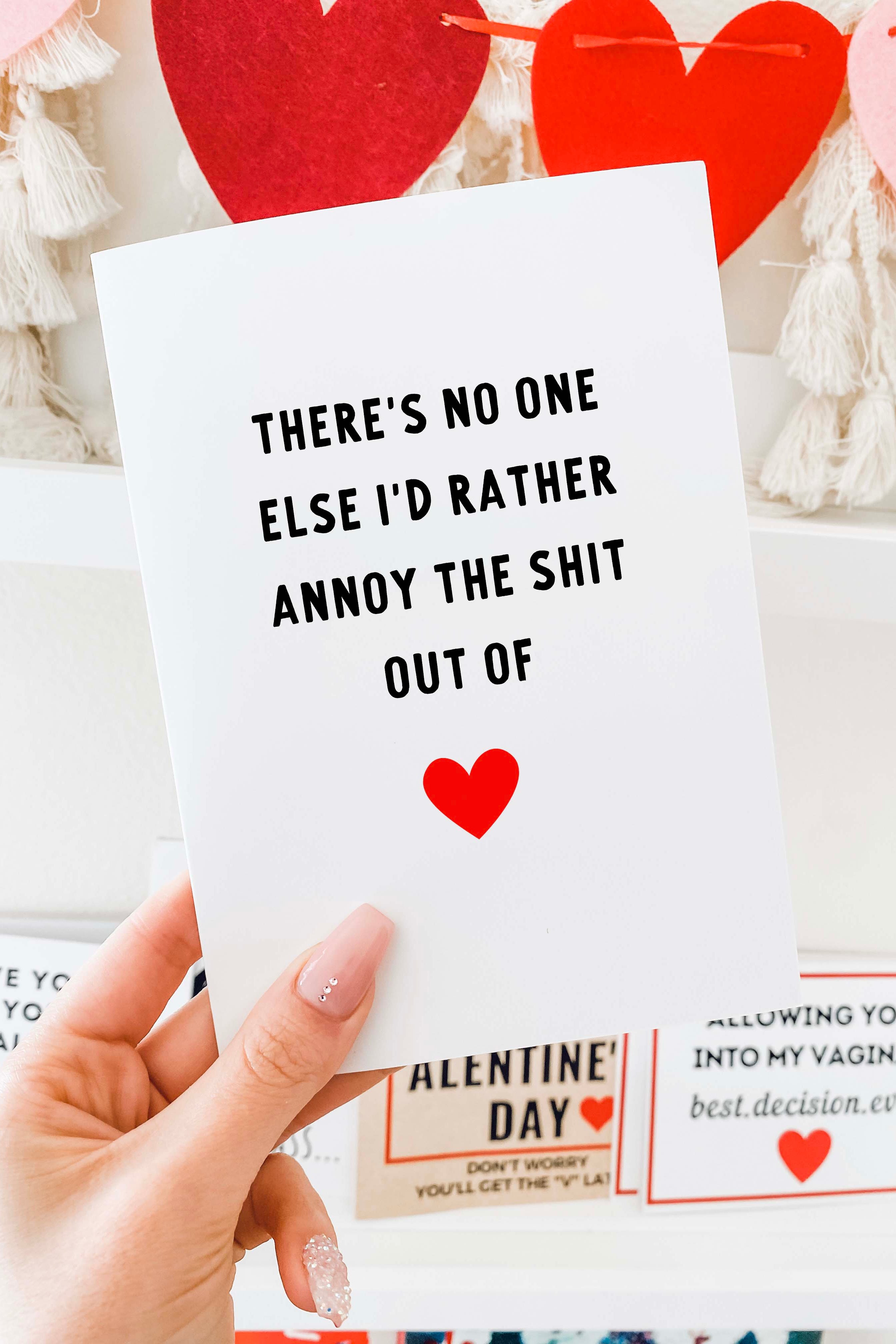 Greeting card with bold black text and a red heart icon, photographed being held by a hand in front of a Valentine themed background