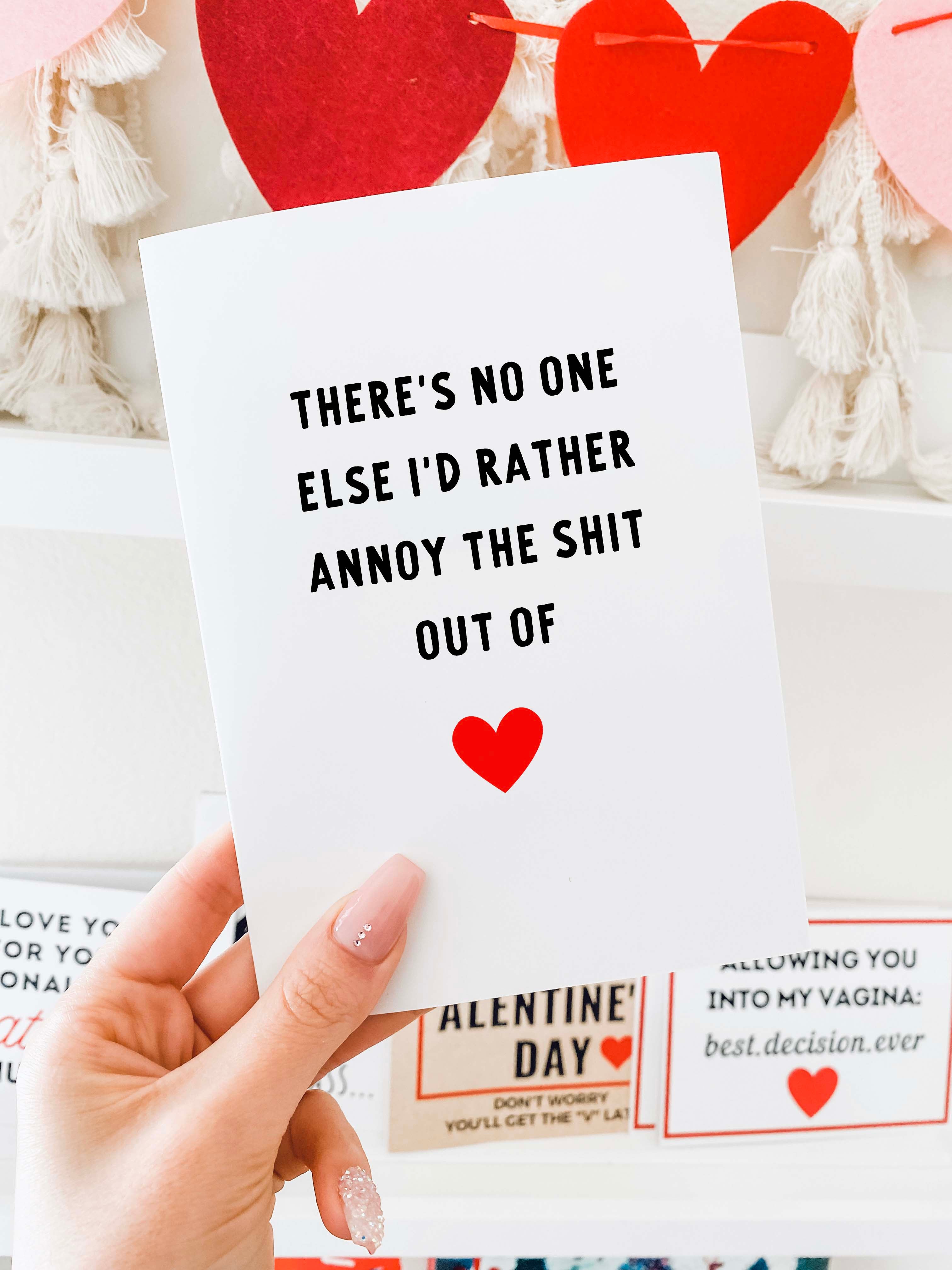 Greeting card with bold black text and a red heart icon, photographed being held by a hand in front of a Valentine themed background