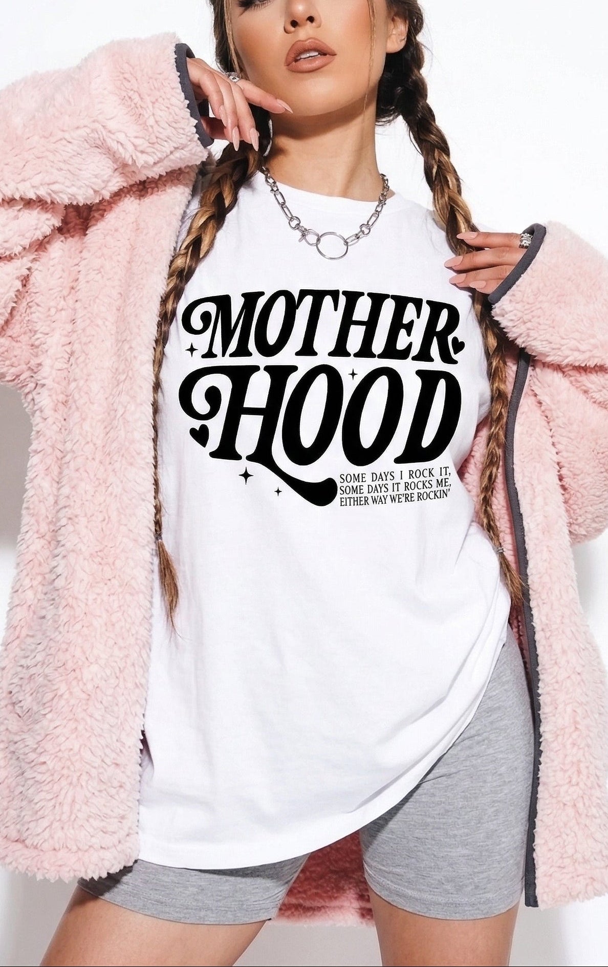 Person wearing a white t-shirt with 'MOTHERHOOD' text, pink fluffy jacket, and gray shorts on a white background