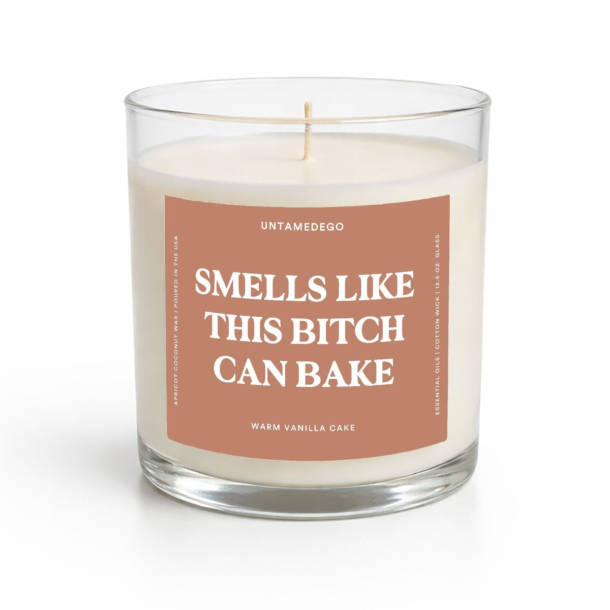 A single-wick soy candle in a clear glass jar with cream-colored wax and a dusty rose label that reads 'SMELLS LIKE THIS BITCH CAN BAKE' by Untamedogo.