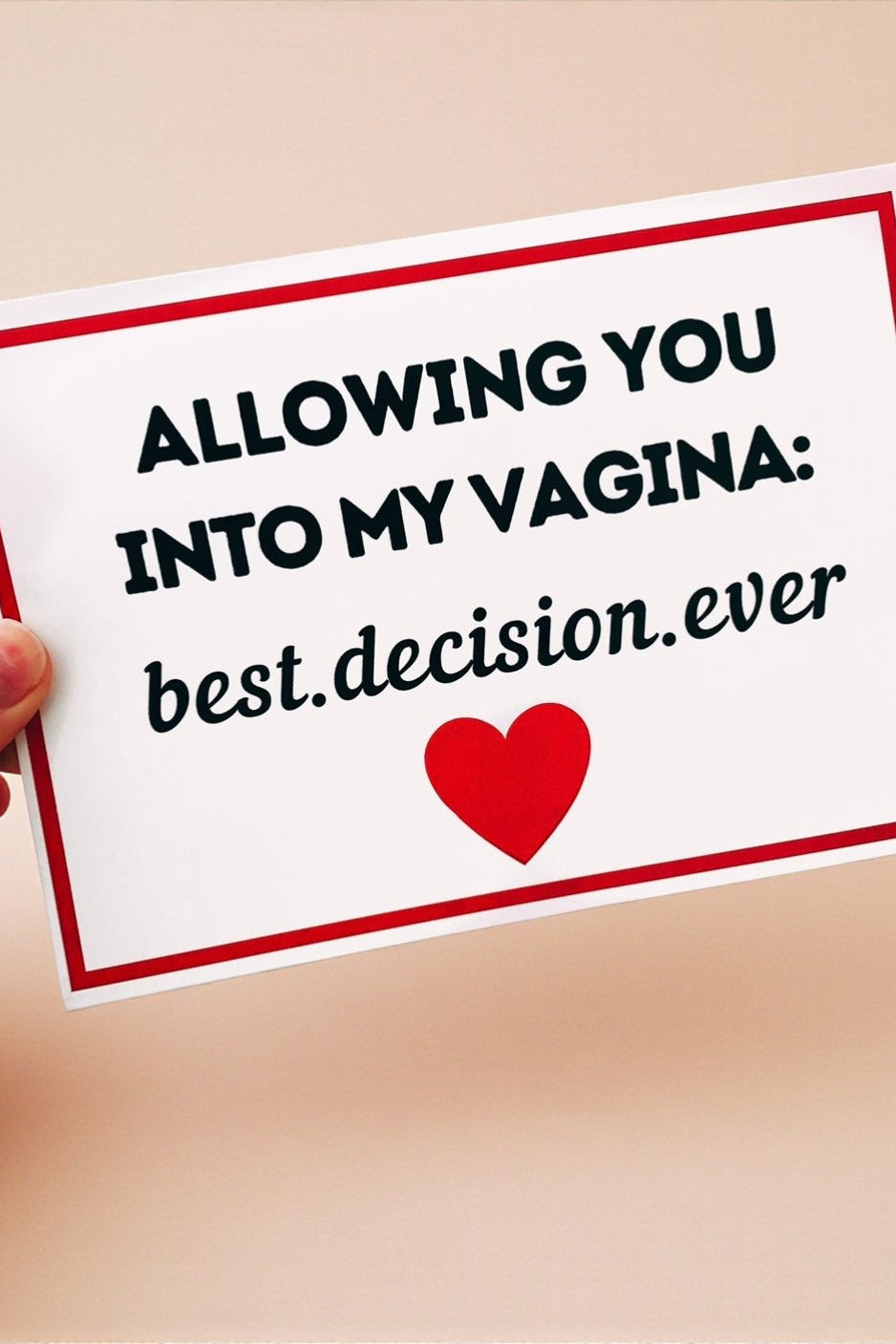 A hand holding a white greeting card with a red border and black text that reads “Allowing you into my vagina: best decision ever,” with a red heart printed below the text. The card is photographed against a warm cream-colored background.