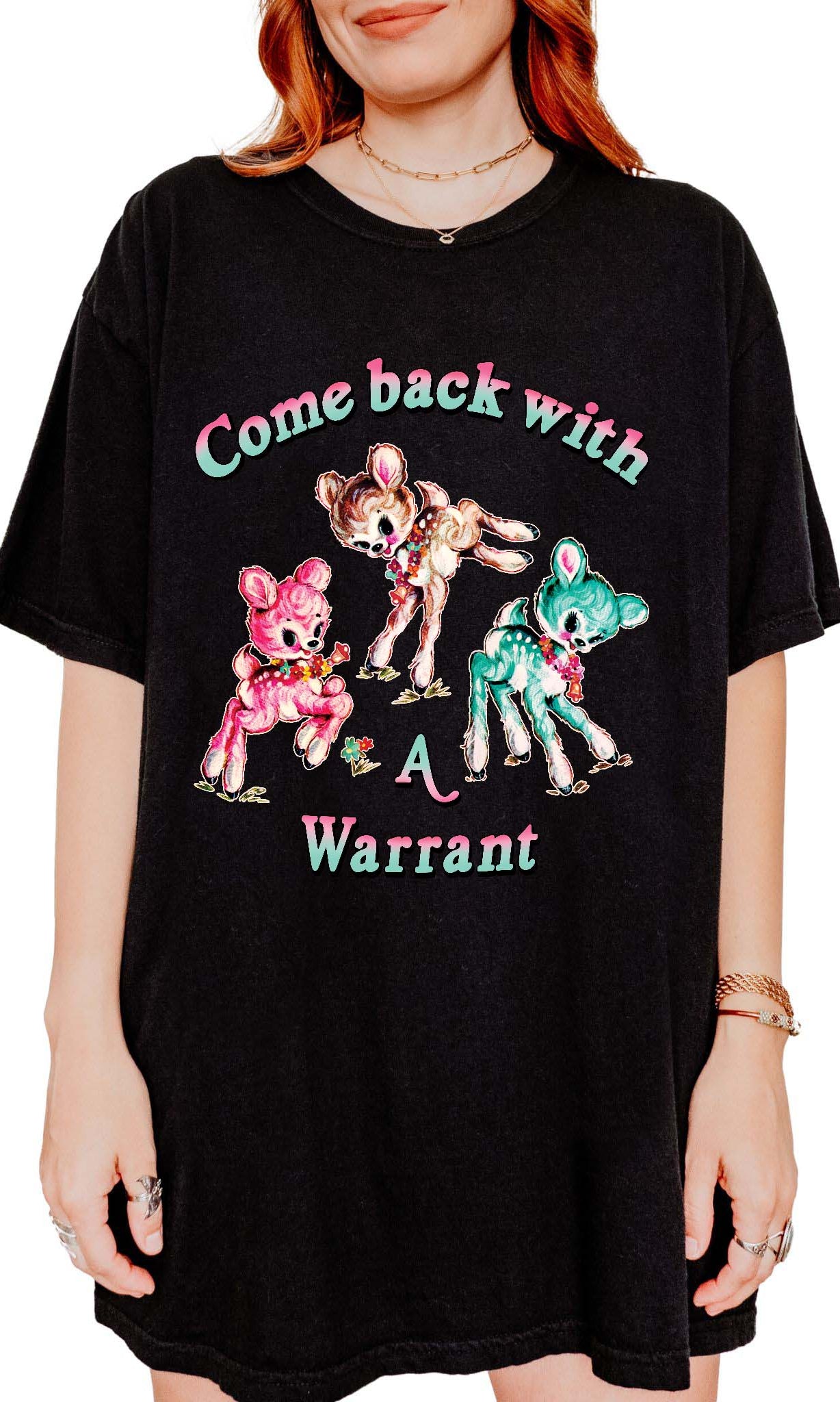 Person wearing a black graphic t-shirt featuring three pastel illustrated fawns and the text Come Back With a Warrant, photographed against a light background