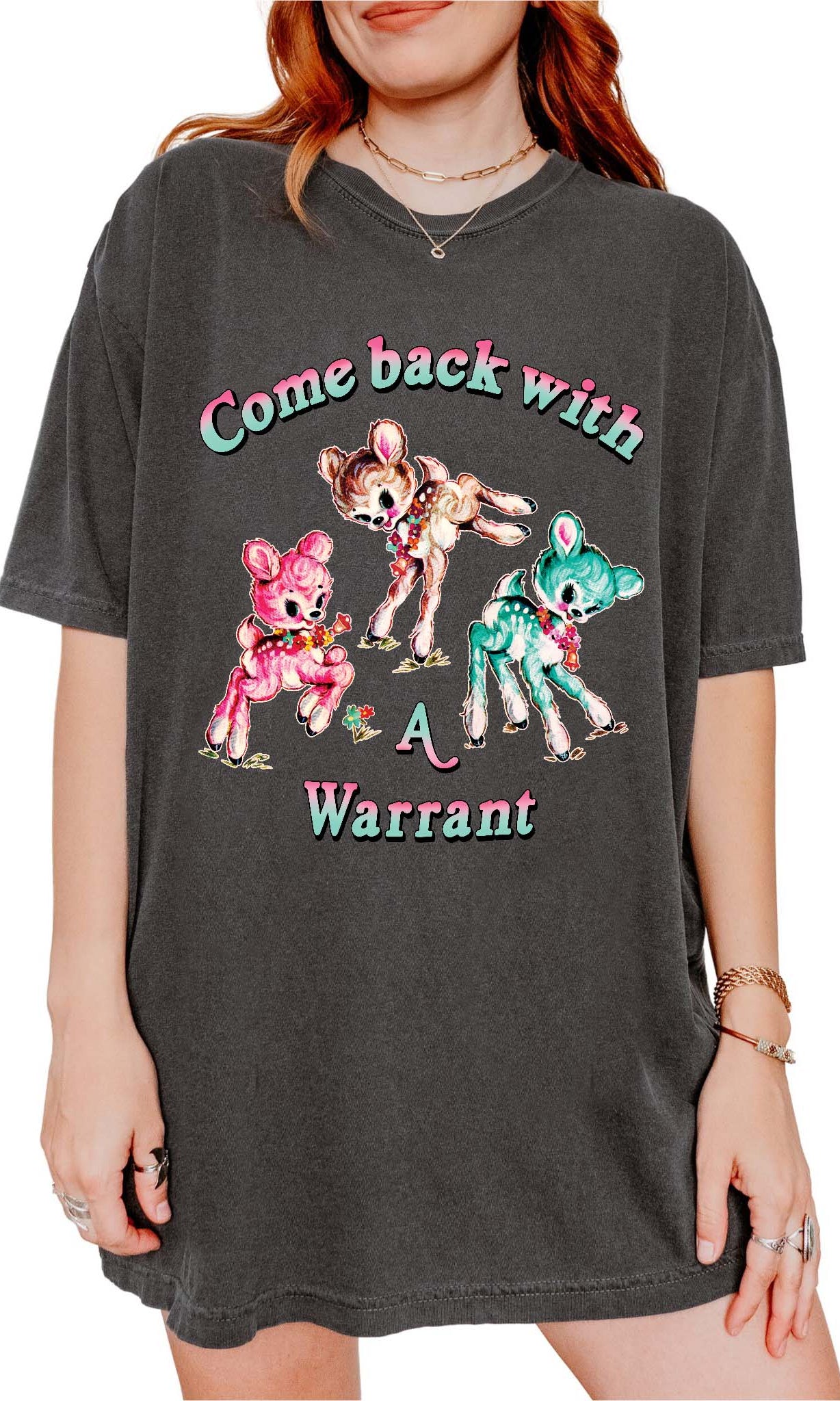 Person wearing a dark gray graphic t-shirt featuring three pastel illustrated fawns and the text Come Back With a Warrant, photographed against a light background