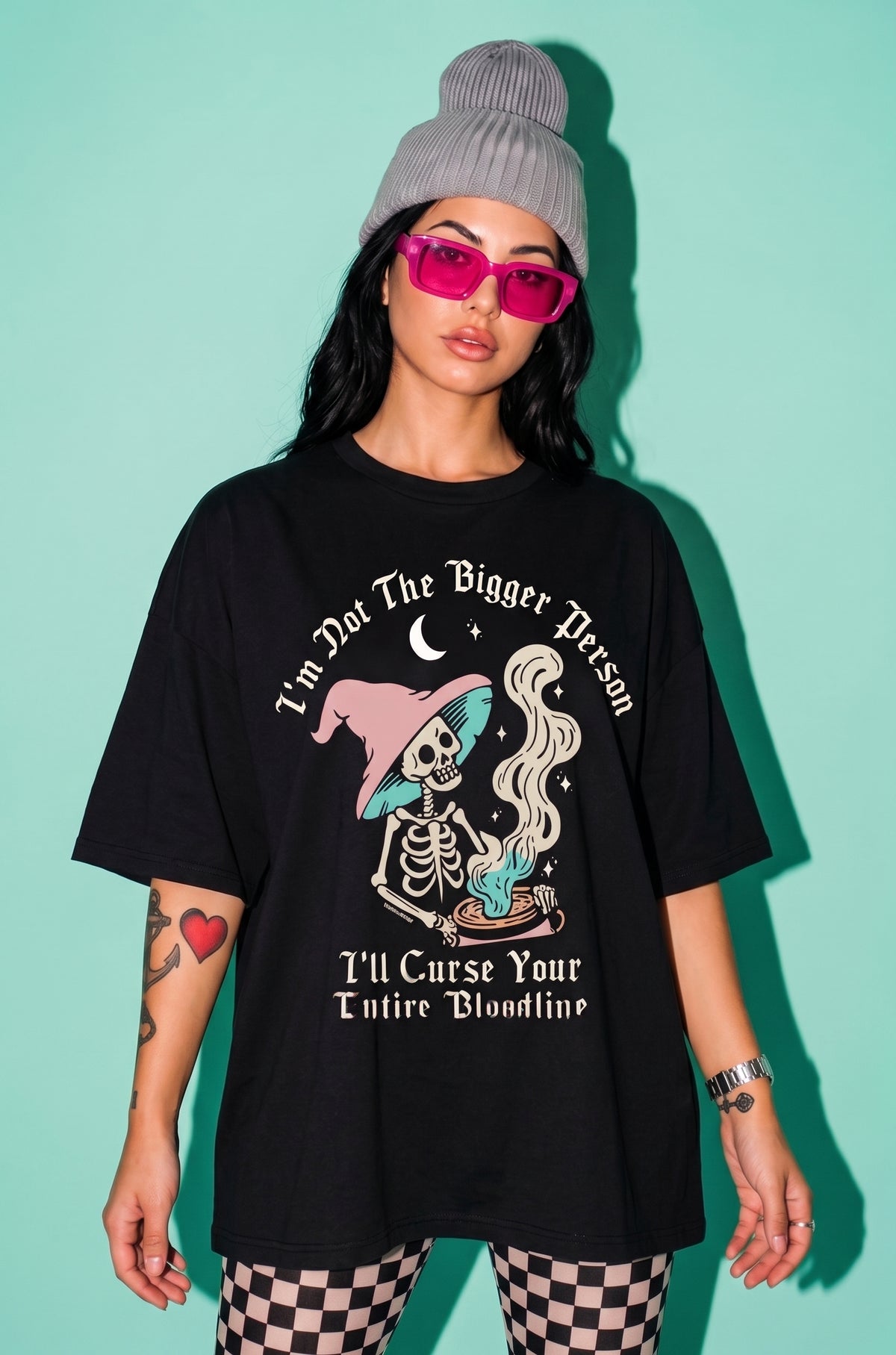 A model wearing a black graphic t-shirt with a skeleton and witch illustration, paired with a gray beanie, pink sunglasses, and checkered leggings, set against a solid mint green background.