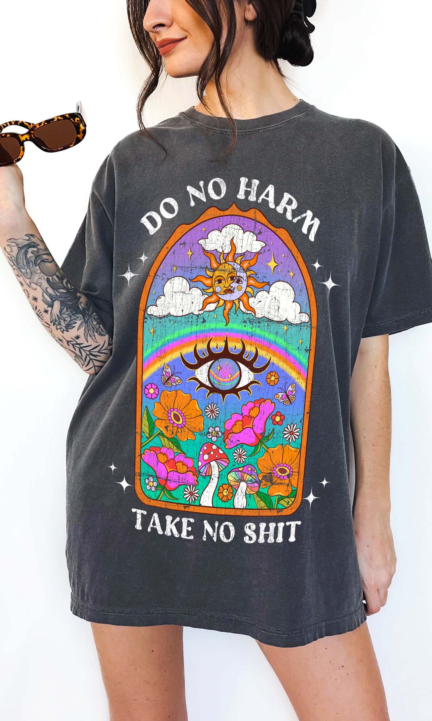 Person wearing a dark gray graphic t-shirt featuring a colorful illustrated scene with sun, rainbow, flowers, mushrooms, and the text Do No Harm Take No Shit, photographed against a light background
