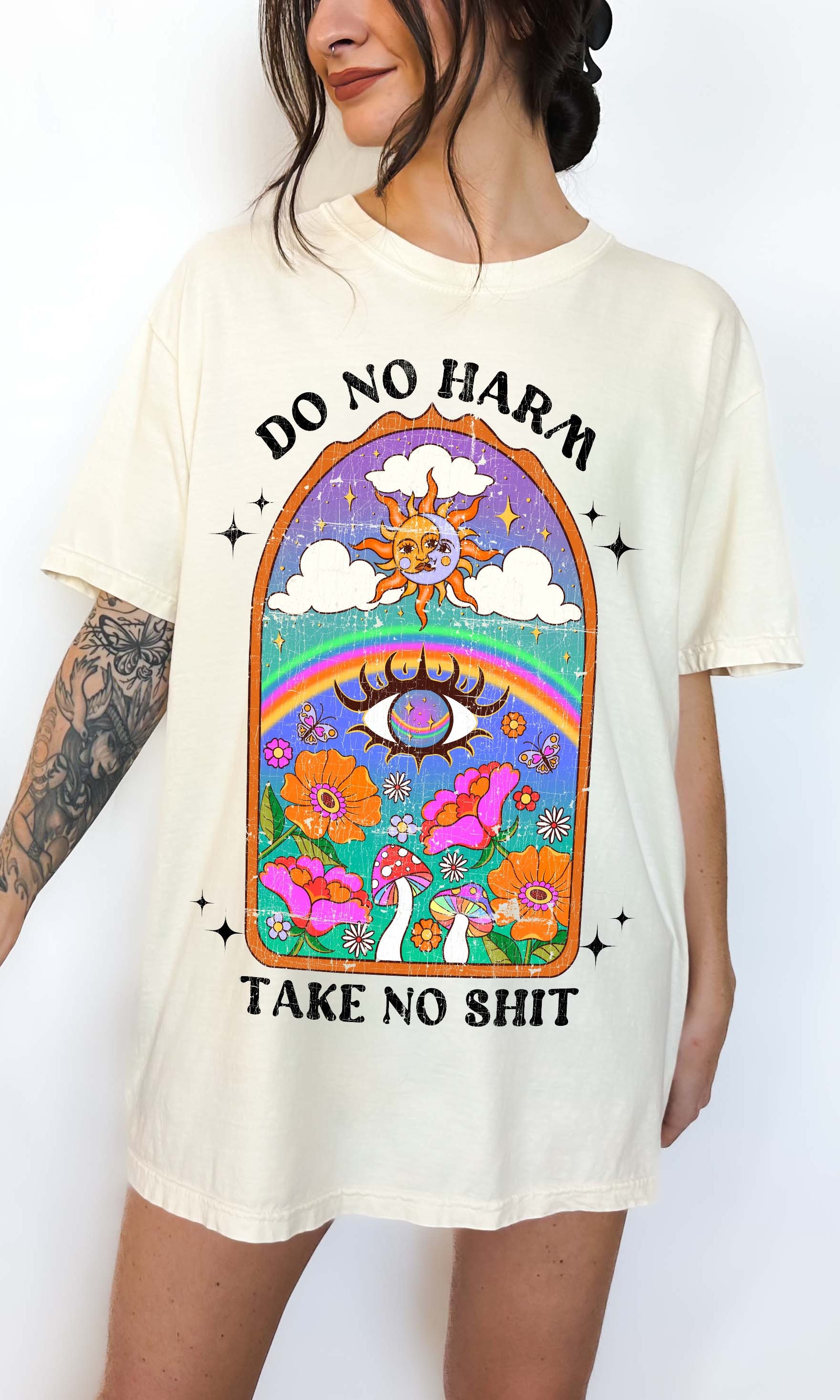 Person wearing Ivory graphic t-shirt featuring a colorful illustrated scene with sun, rainbow, flowers, mushrooms, and the text Do No Harm Take No Shit, photographed against a light background
