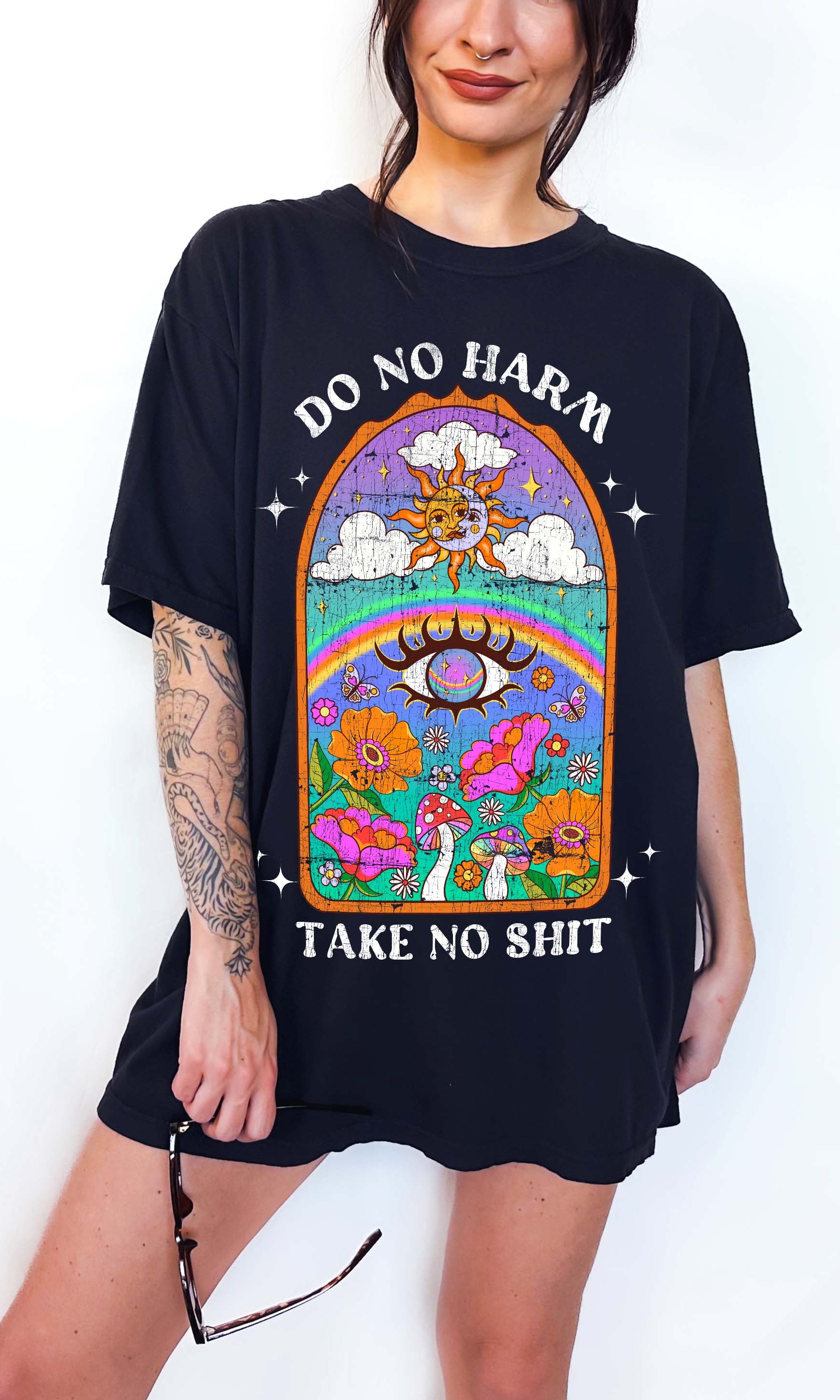 Person wearing a black graphic t-shirt featuring a colorful illustrated scene with sun, rainbow, flowers, mushrooms, and the text Do No Harm Take No Shit, photographed against a light background