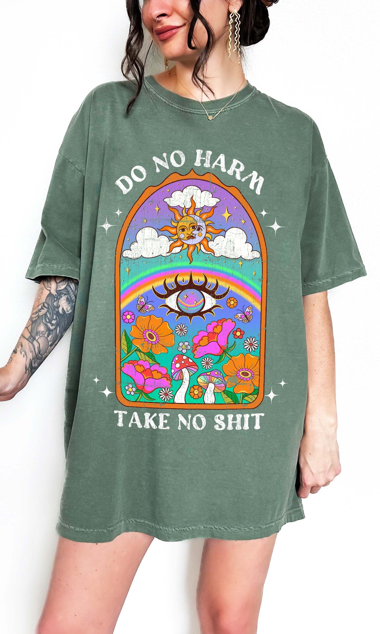 Person wearing a green graphic t-shirt featuring a colorful illustrated scene with sun, rainbow, flowers, mushrooms, and the text Do No Harm Take No Shit, photographed against a light background