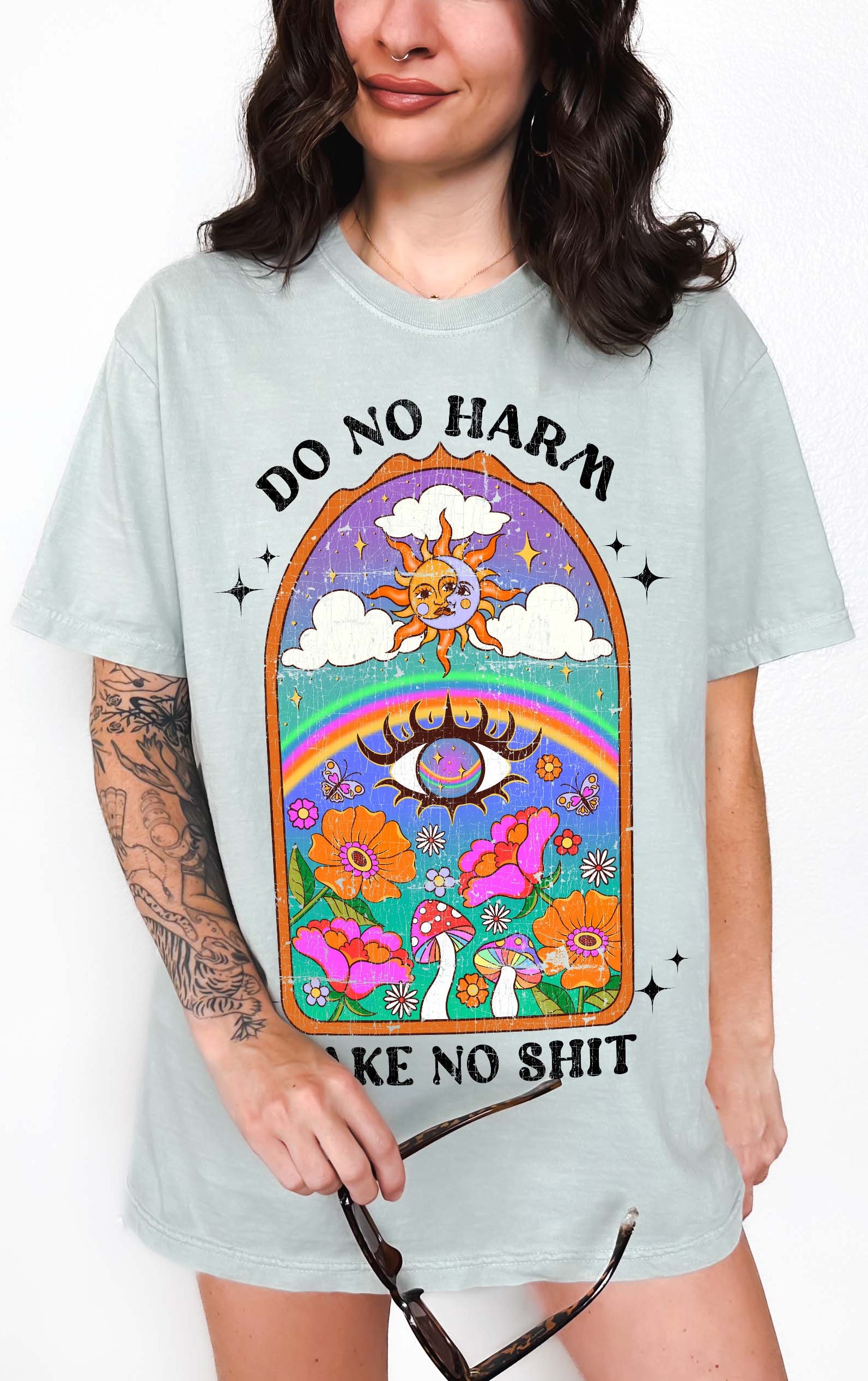 Person wearing a light blue graphic t-shirt featuring a colorful illustrated scene with sun, rainbow, flowers, mushrooms, and the text Do No Harm Take No Shit, photographed against a light background