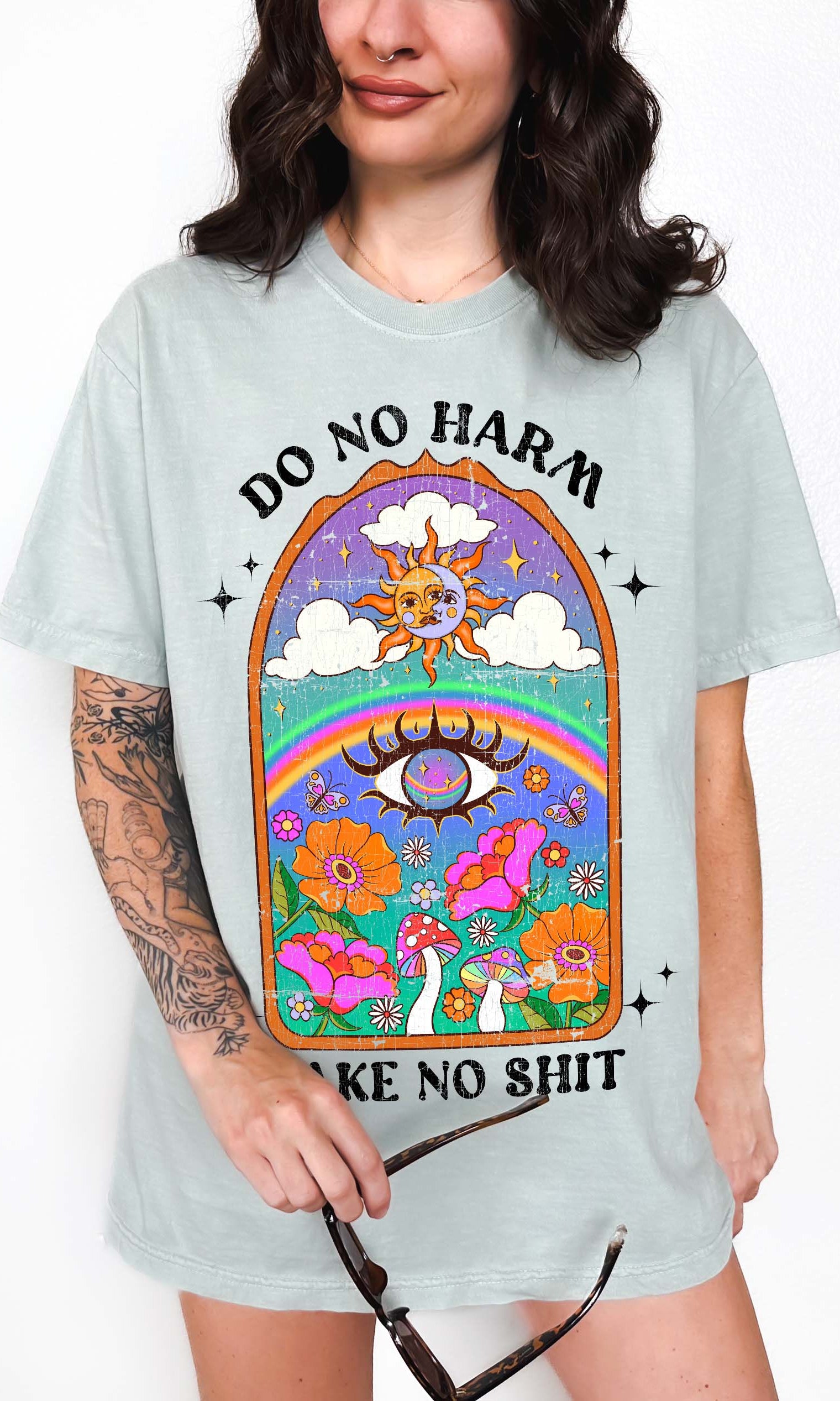Person wearing a light blue graphic t-shirt featuring a colorful illustrated scene with sun, rainbow, flowers, mushrooms, and the text Do No Harm Take No Shit, photographed against a light background