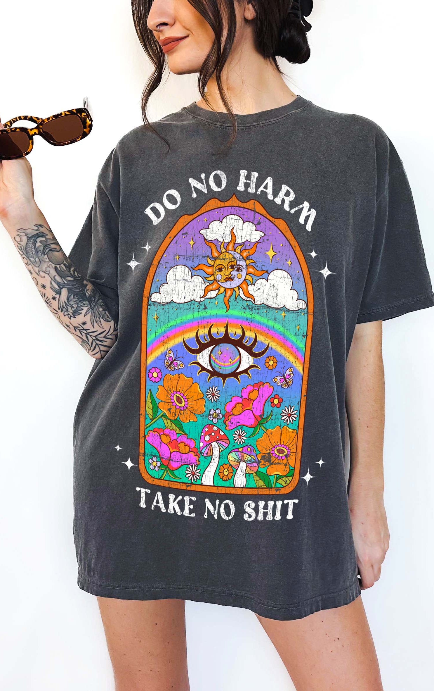 Person wearing a dark gray graphic t-shirt featuring a colorful illustrated scene with sun, rainbow, flowers, mushrooms, and the text Do No Harm Take No Shit, photographed against a light background