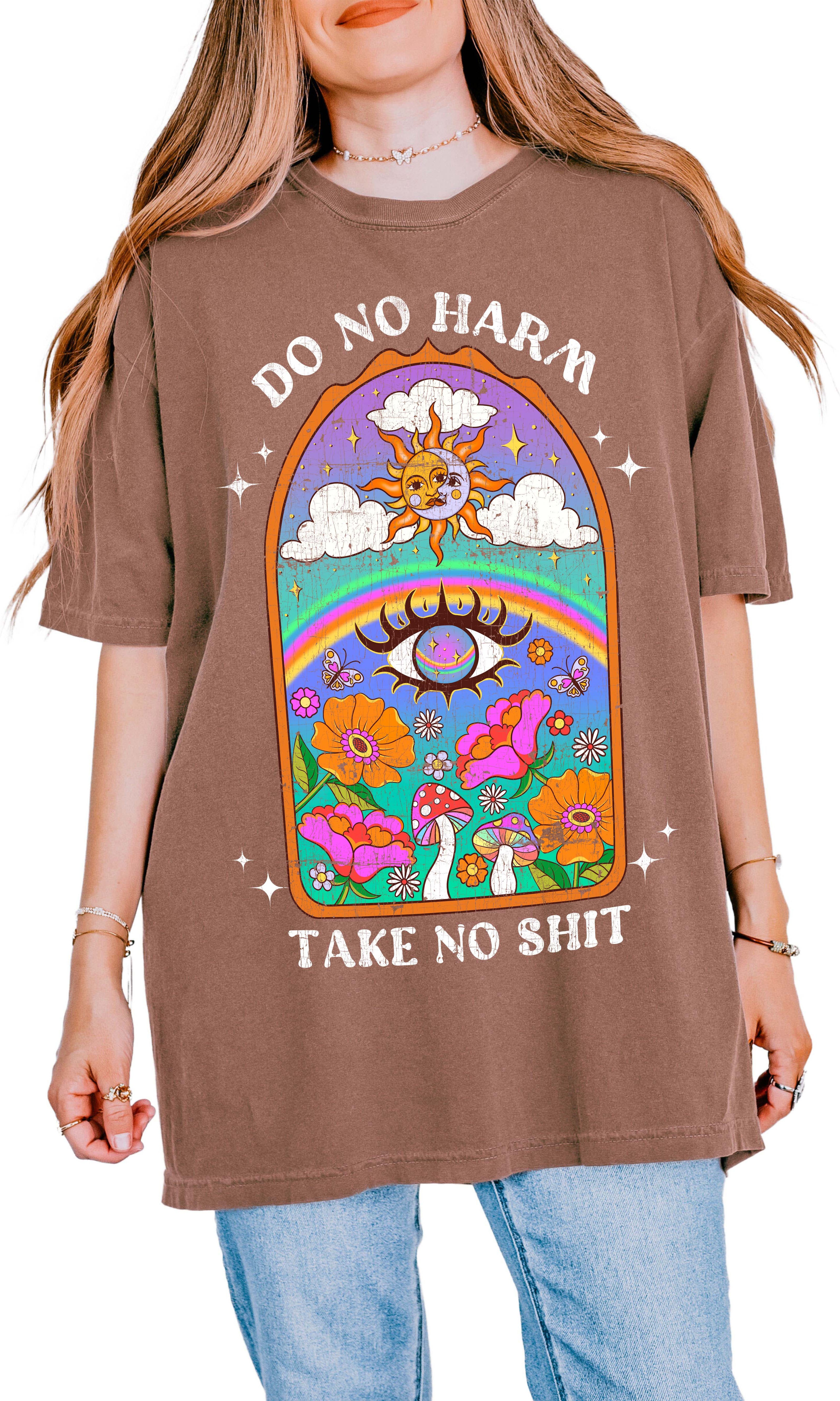 Person wearing a brown graphic t-shirt featuring a colorful illustrated scene with sun, rainbow, flowers, mushrooms, and the text Do No Harm Take No Shit, photographed against a light background