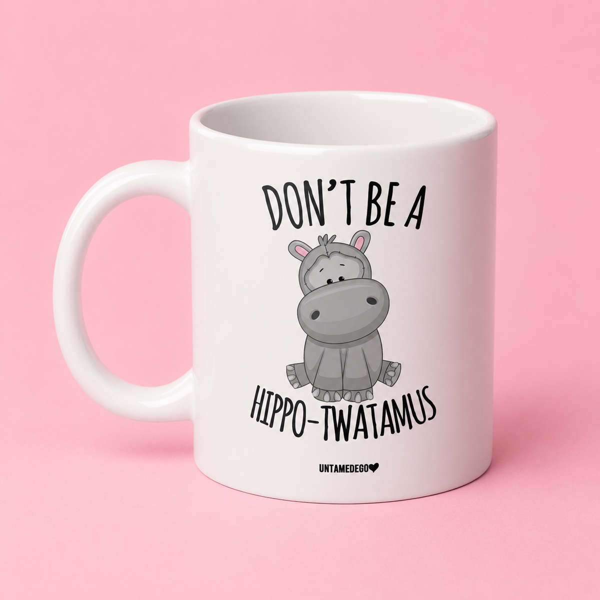 A white ceramic mug with a cartoon hippo graphic and the text 'DON'T BE A HIPPO-TWATAMUS' printed on it, set against a solid pink background.