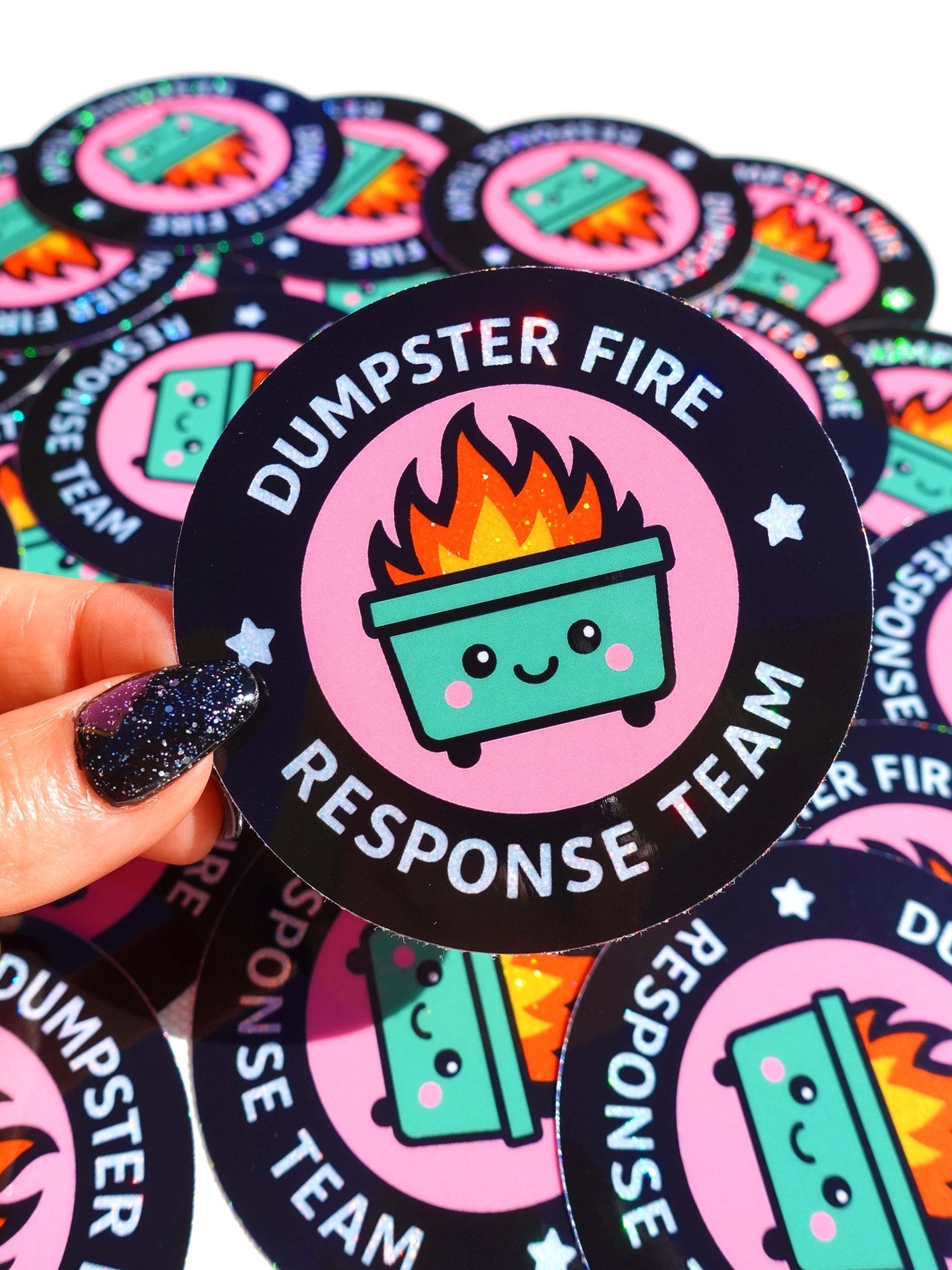Round holographic glitter sticker reading "Dumpster Fire Response Team" held by a hand with dark glitter nails against a yellow background, surrounded by multiples of the same sticker on a dark surface.