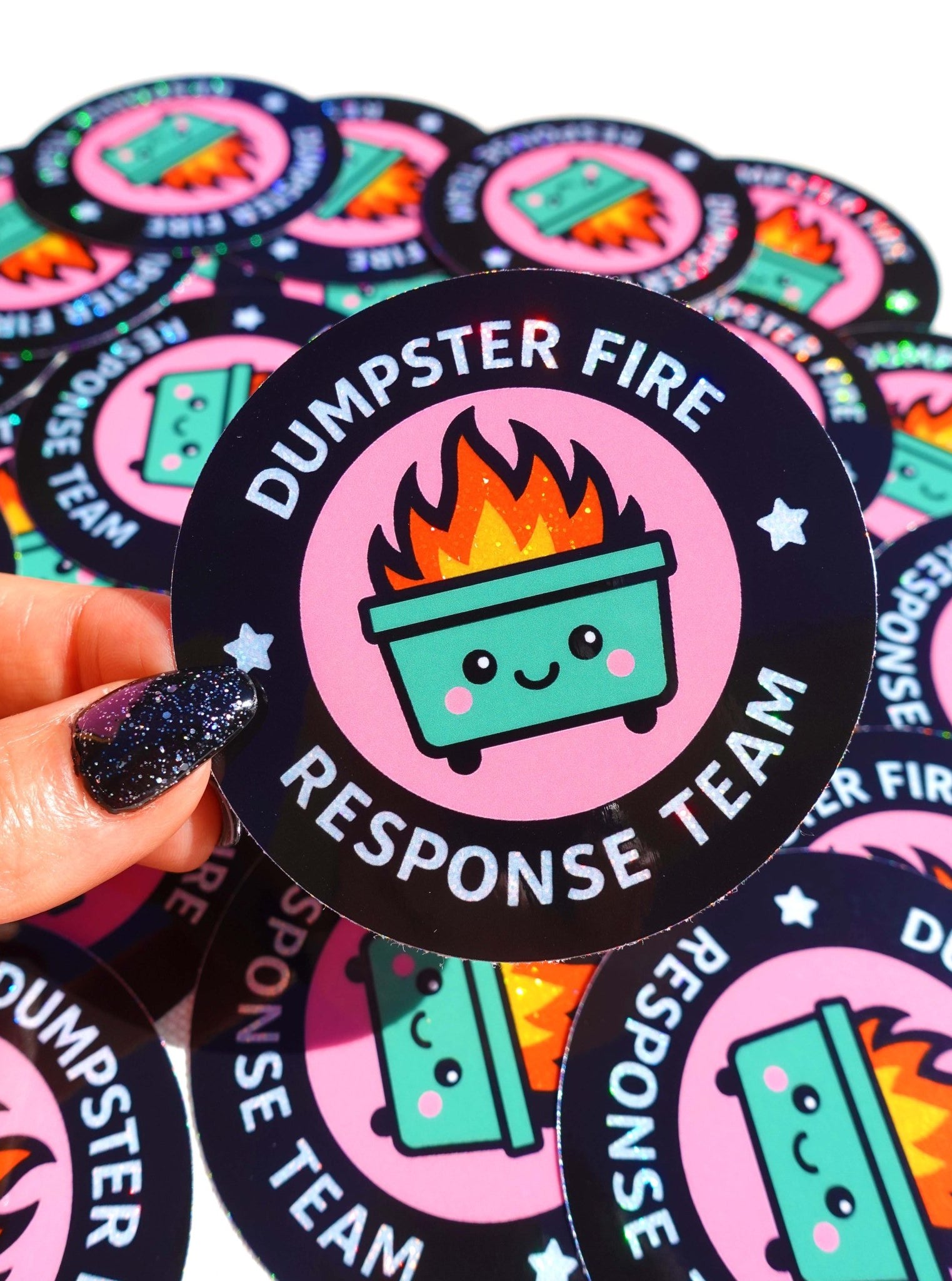 Round holographic glitter sticker reading "Dumpster Fire Response Team" held by a hand with dark glitter nails against a yellow background, surrounded by multiples of the same sticker on a dark surface.