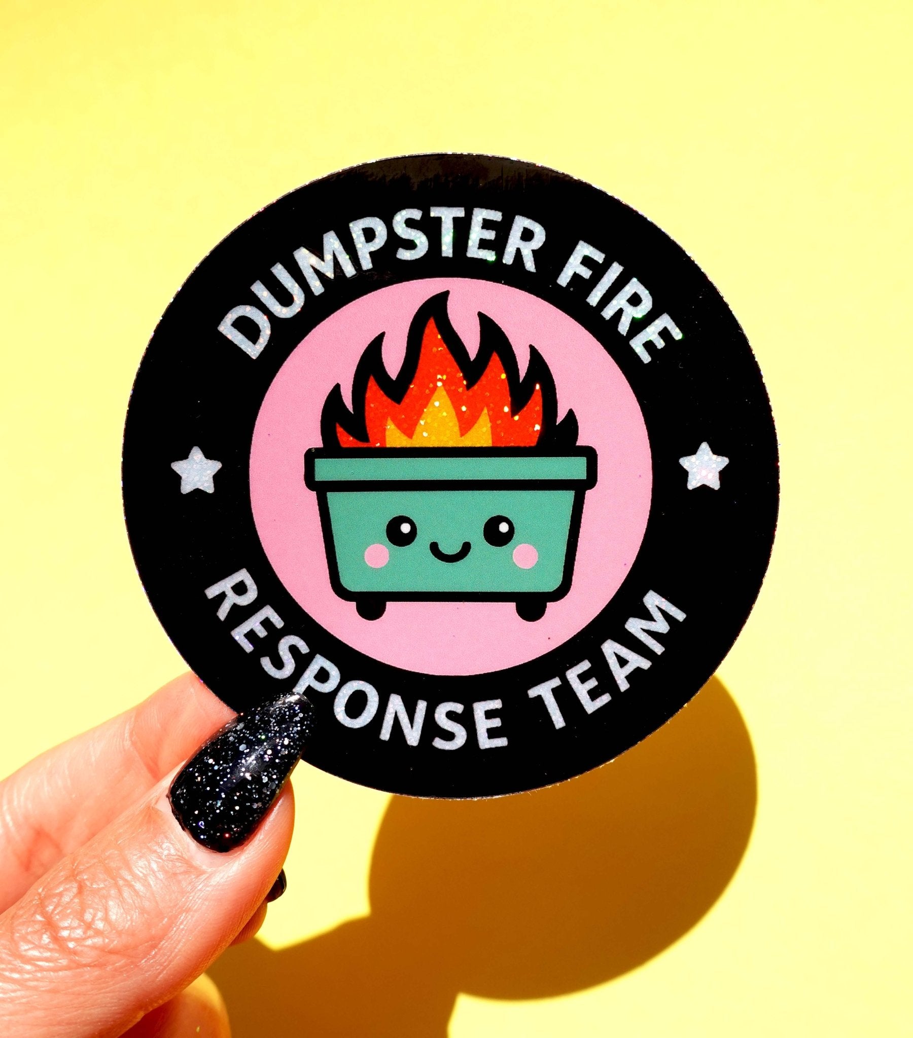 Round die-cut vinyl sticker with a black background and pink circle featuring a kawaii green dumpster with flames and a smiley face. White arched text reads Dumpster Fire Response Team with star accents.