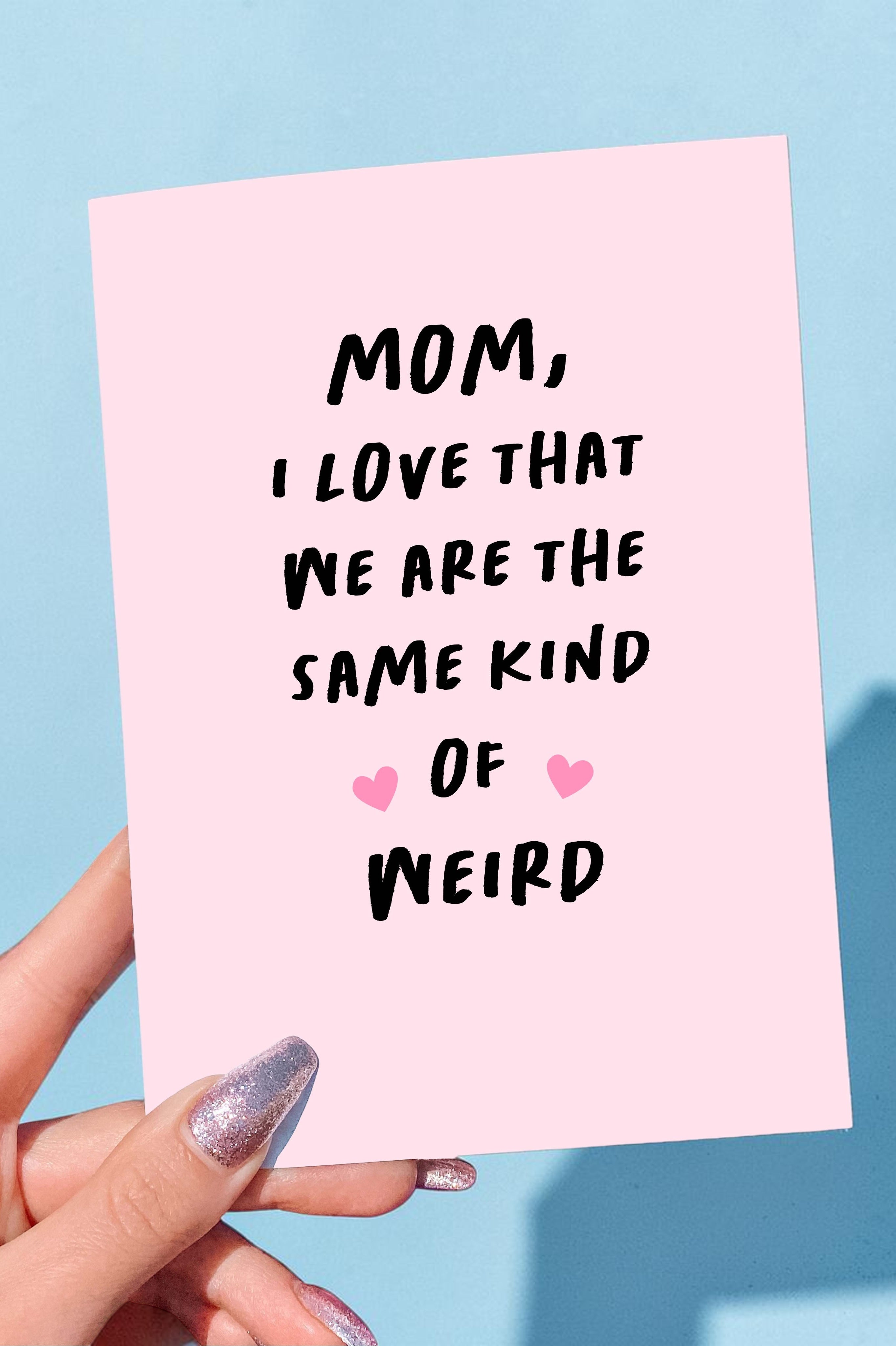 Pink card with text 'Mom, I love that we are the same kind of weird' held by a hand against a blue background