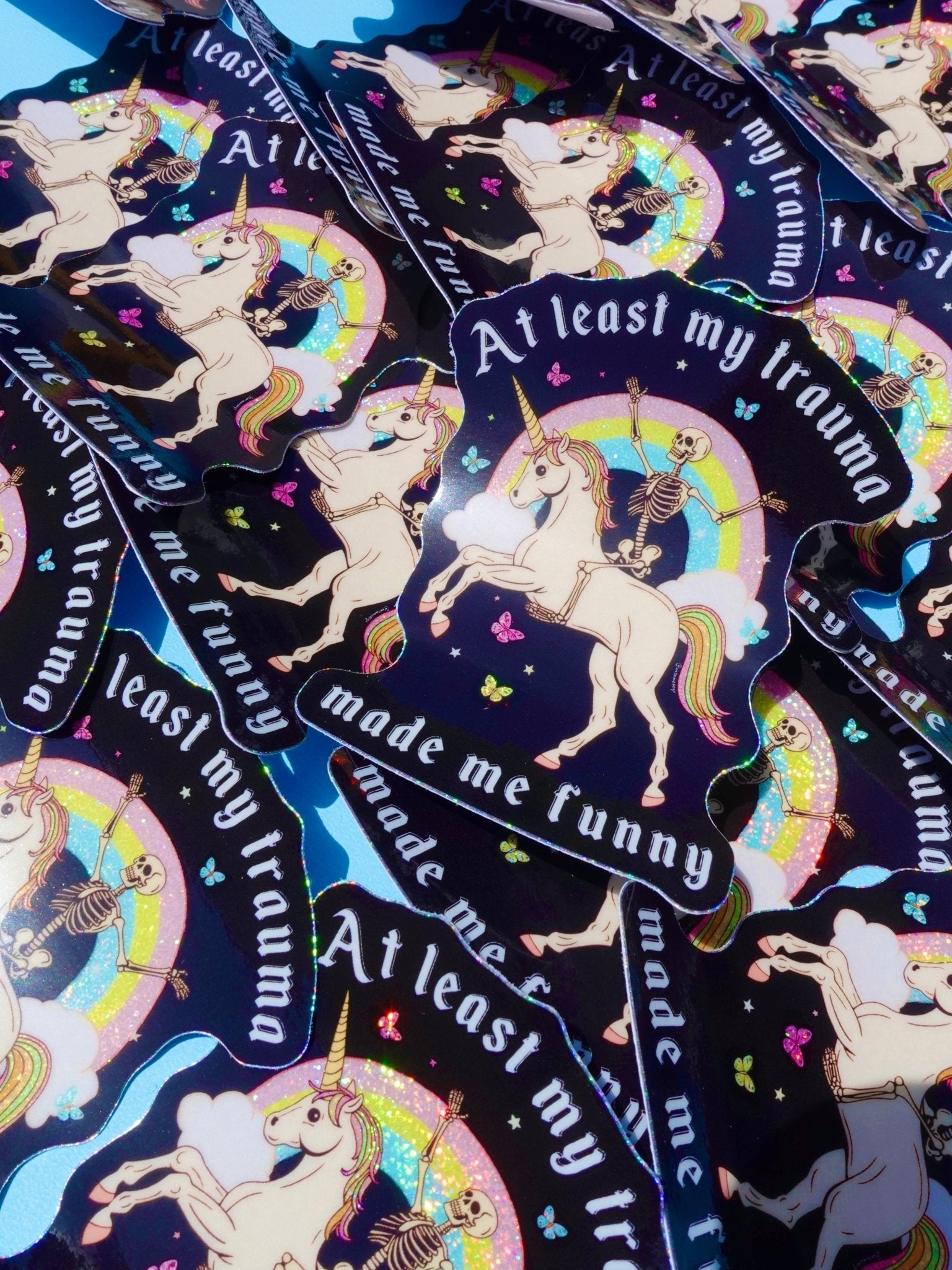 Holographic glitter vinyl stickers showing a skeleton riding a unicorn under a rainbow with butterflies and clouds. Bold white text reads At Least My Trauma Made Me Funny.