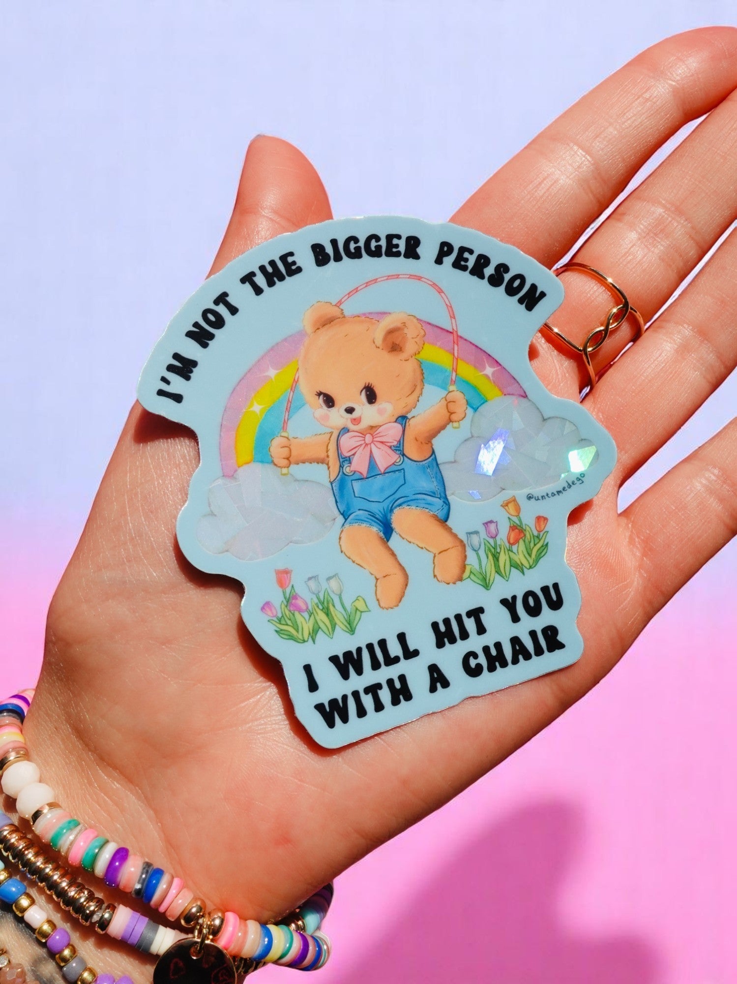 Holographic glitter vinyl bear sticker reading I'm Not The Bigger Person I Will Hit You With A Chair, shown in hand against pink background.