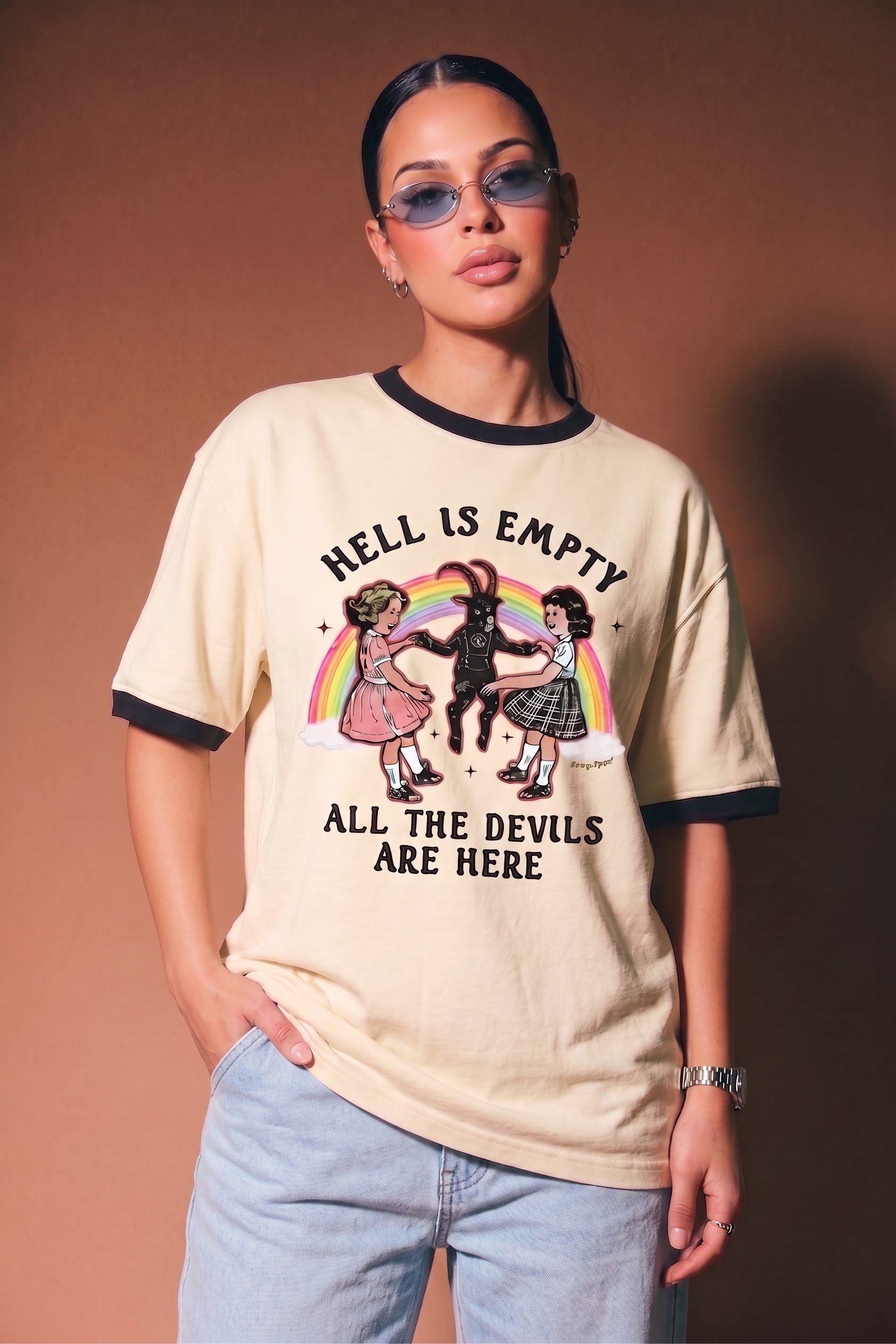 A woman modeling a beige ringer t-shirt with black trim and a graphic print featuring a rainbow, three figures, and the text 'HELL IS EMPTY ALL THE DEVILS ARE HERE'. She is wearing light blue jeans against a solid brown studio background.