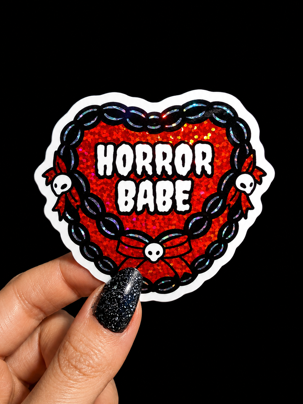 A hand with glittery black nail polish holds a die-cut sticker featuring a red glitter heart, black chain border, white skulls, and the text 'HORROR BABE' in white. The background is solid black.