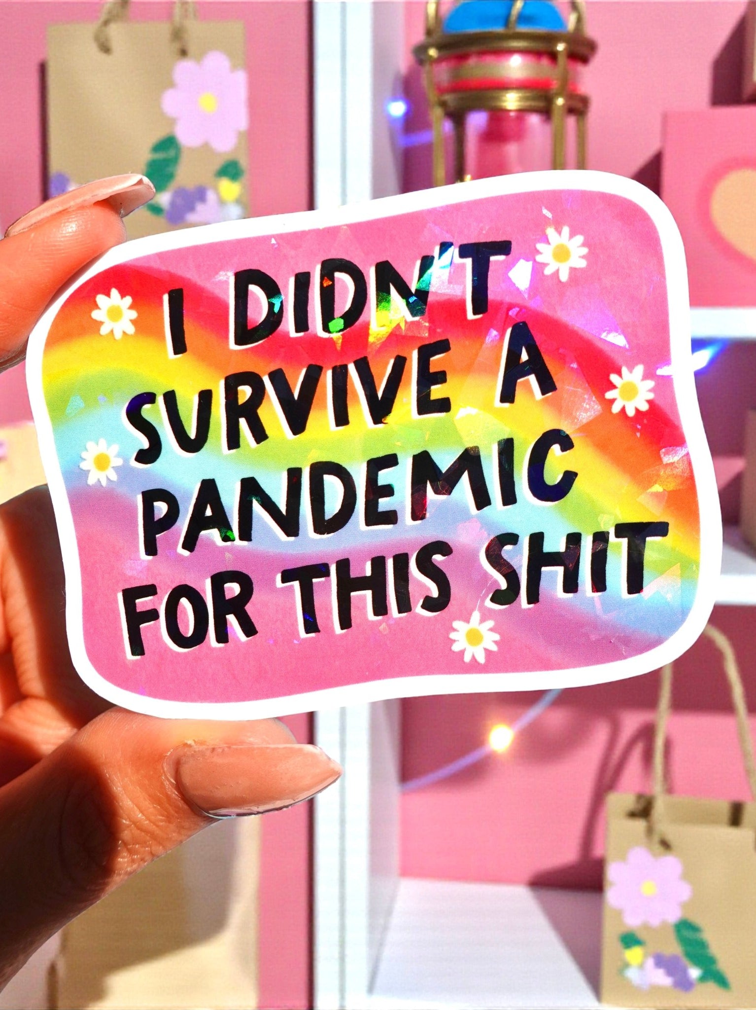 Rounded rectangle die-cut holographic vinyl sticker with a rainbow swirl background and white daisy accents along the border. Bold black text reads I Didn't Survive a Pandemic for This Shit.