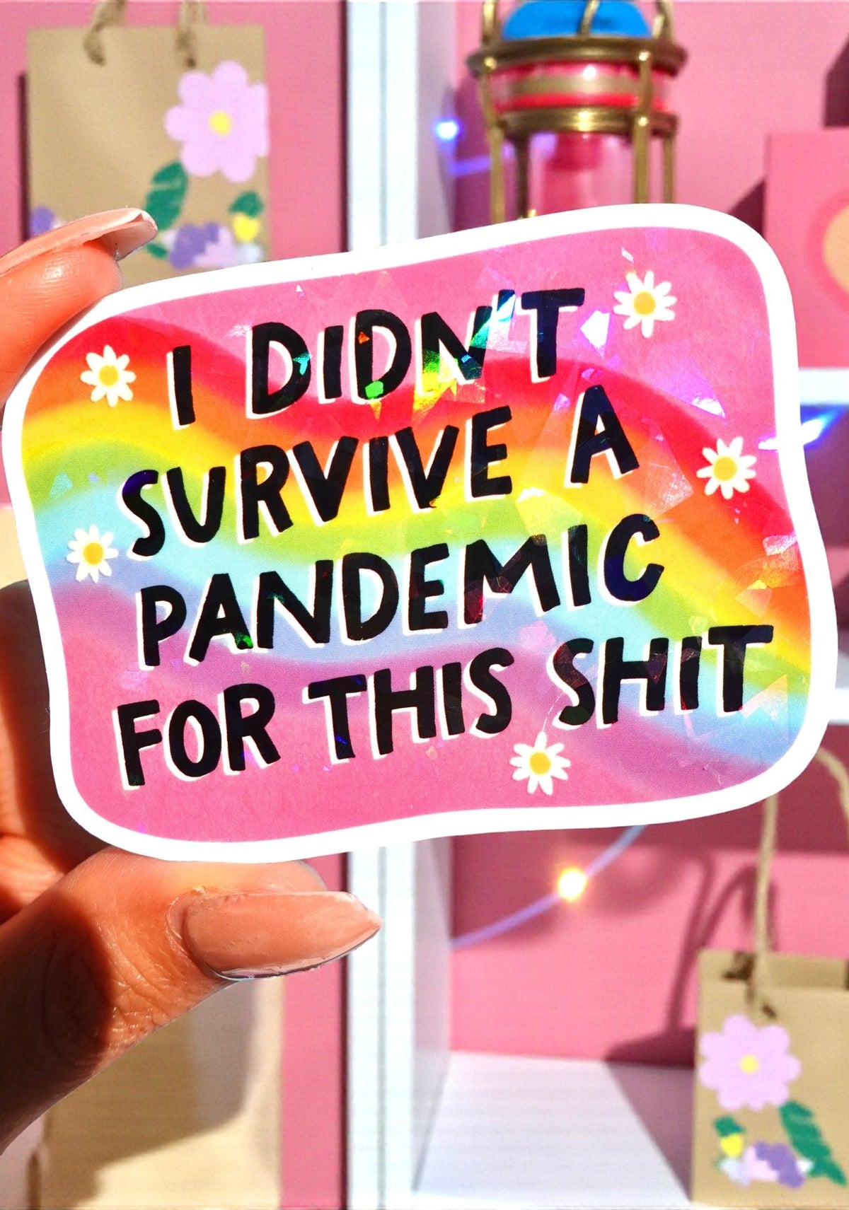 Rounded rectangle die-cut holographic vinyl sticker with a rainbow swirl background and white daisy accents along the border. Bold black text reads I Didn't Survive a Pandemic for This Shit.