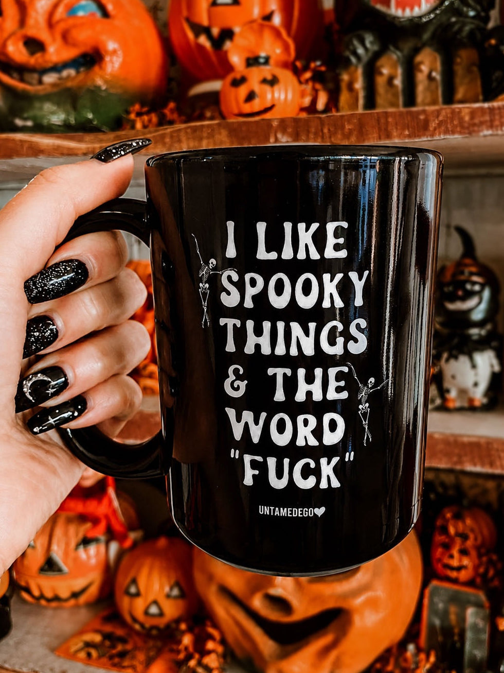 A hand with glittery black nails holds a black ceramic mug with white text that reads 'I LIKE SPOOKY THINGS & THE WORD "FUCK"' and the brand name 'UNTAMEDGO' at the bottom. The background shows shelves filled with Halloween decorations, including pumpkins and a spooky skull figure.