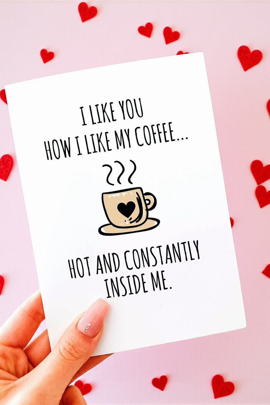 A hand holding a white greeting card with black text that reads “I like you how I like my coffee… hot and constantly inside me,” with a small illustrated coffee cup featuring a heart. The background is soft blush pink with scattered red heart shapes.
