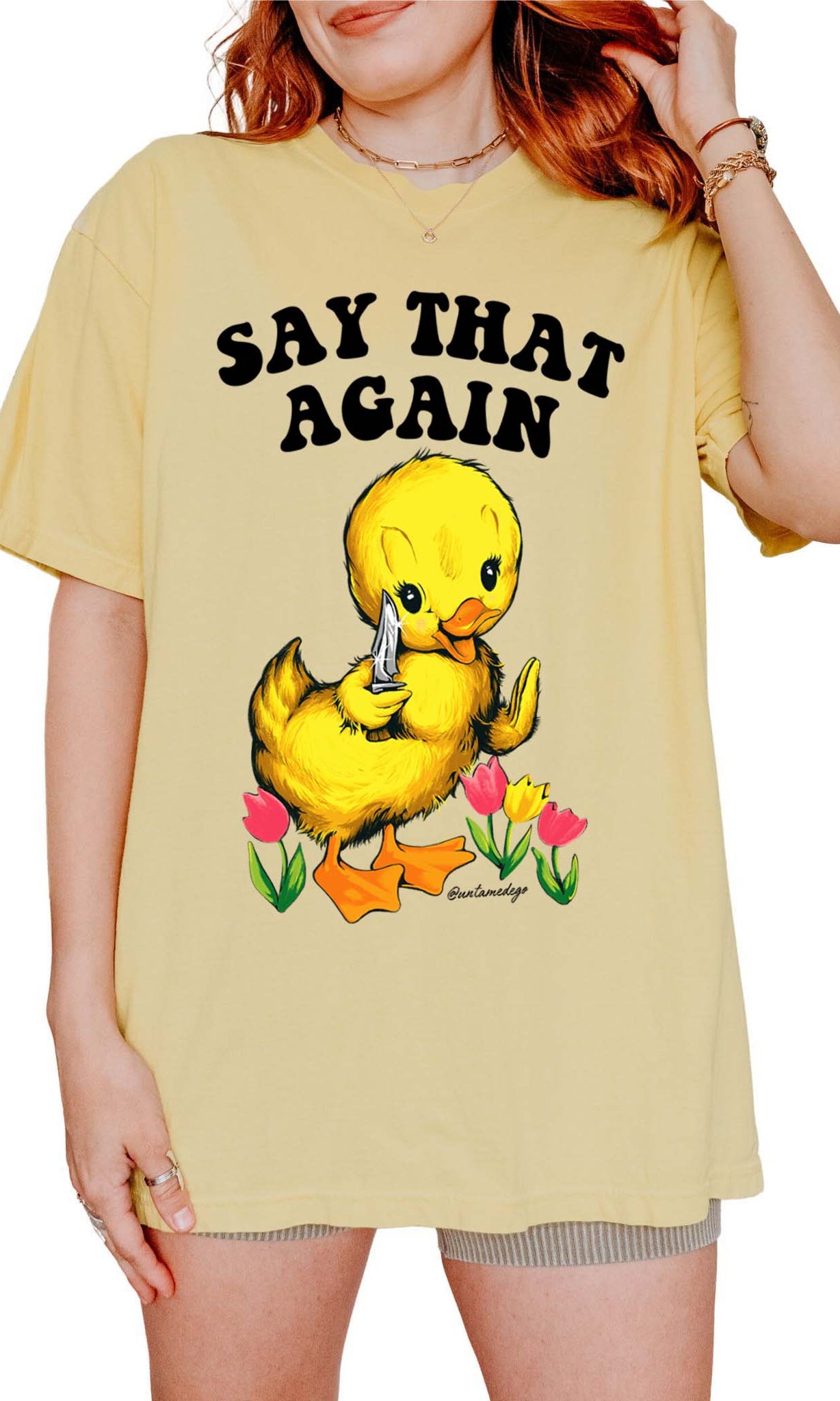 A person wearing a yellow crew neck t-shirt with a graphic that says 'SAY THAT AGAIN' and features an illustration of a duck with a flower. The t-shirt has a relaxed fit.
