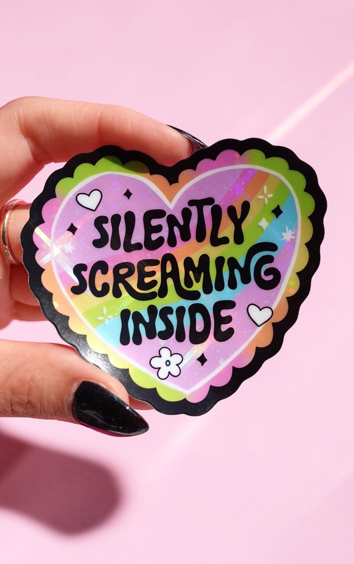 Holographic heart sticker reading "Silently Screaming Inside" held by a hand with dark nails against a pink background, showing pastel rainbow colors and black scalloped border detail.