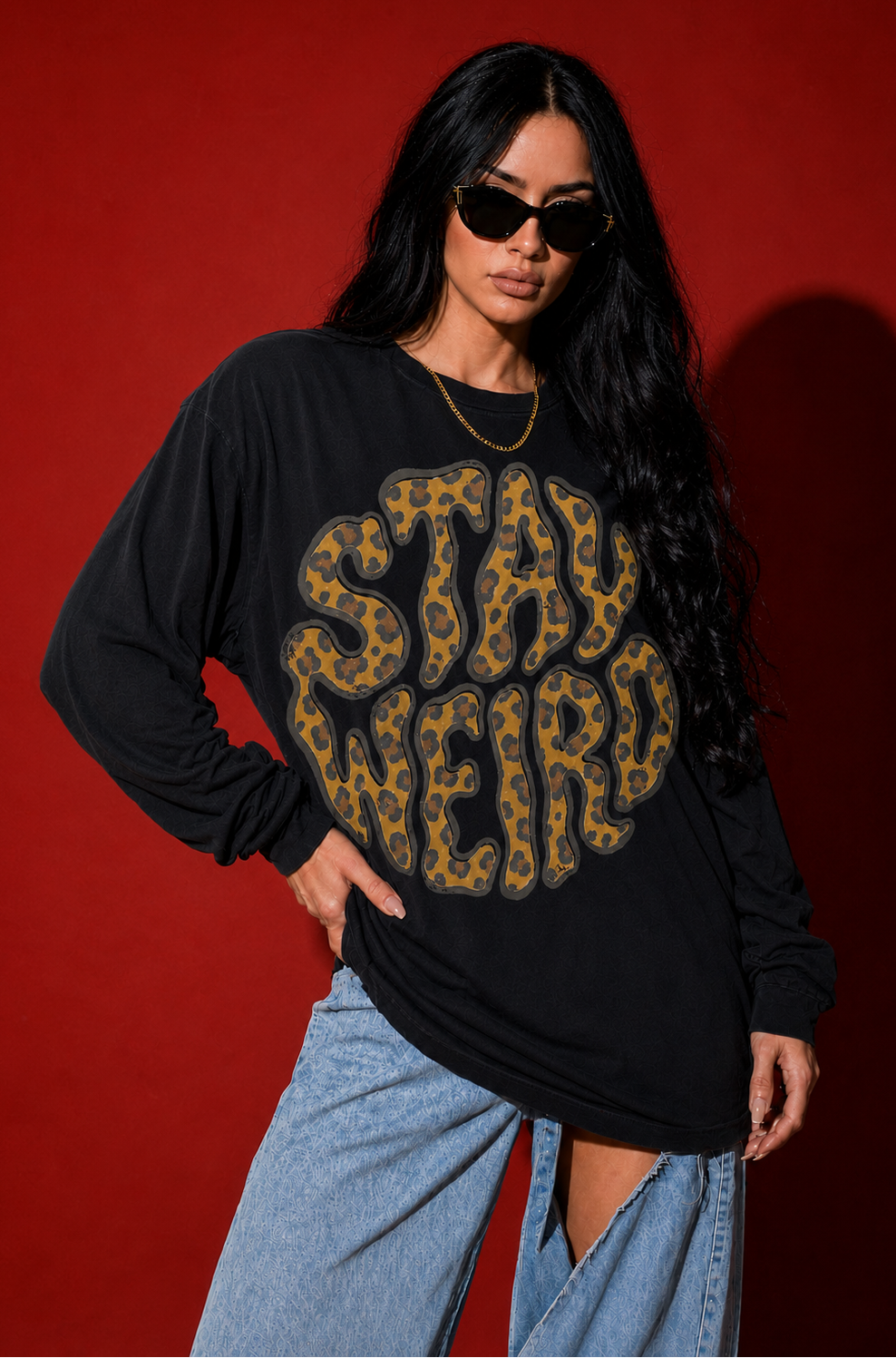 A woman modeling a black long-sleeve graphic T-shirt with a large leopard-print 'STAY WEIRD' design, paired with light blue jeans, against a solid red studio background.