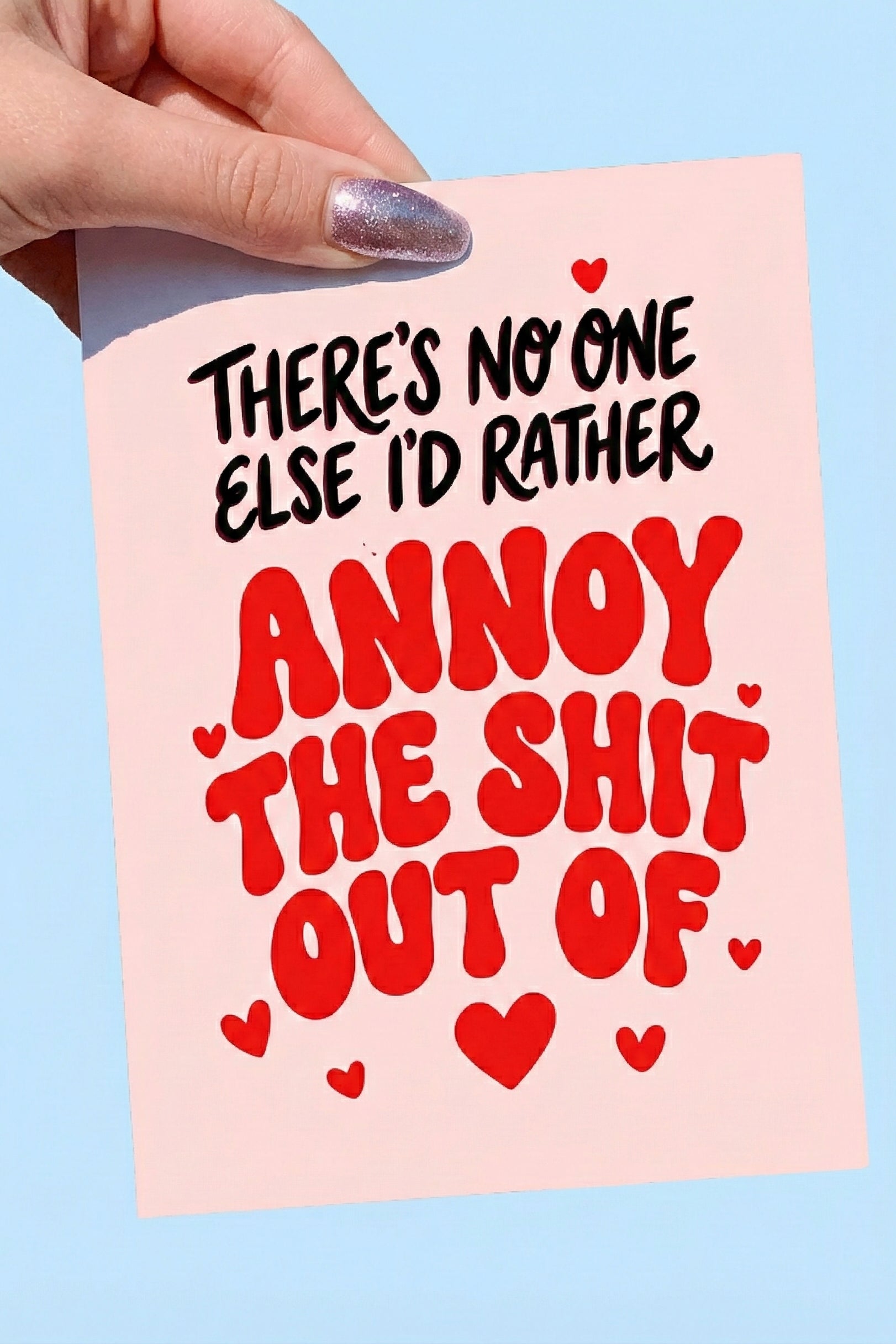Greeting card with bold red and black text and heart graphics on a pink background, photographed being held by a hand