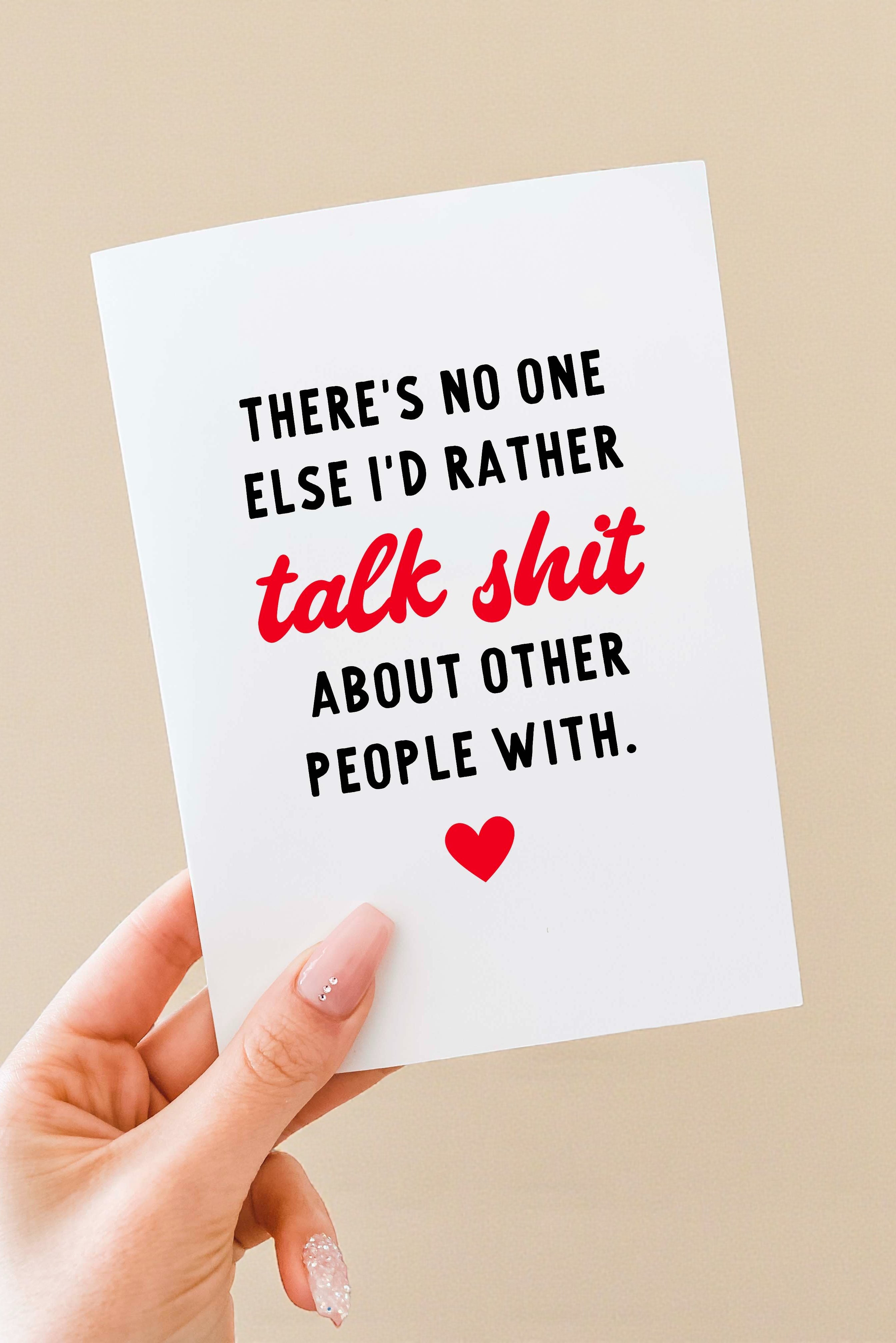 Greeting card held in a hand against a neutral background. The card has a white background with bold black text that reads, “There’s no one else I’d rather,” followed by the phrase “talk shit” in red script-style lettering, and then black text reading, “about other people with.” A small red heart icon appears centered near the bottom of the card. The card is vertically oriented and blank inside
