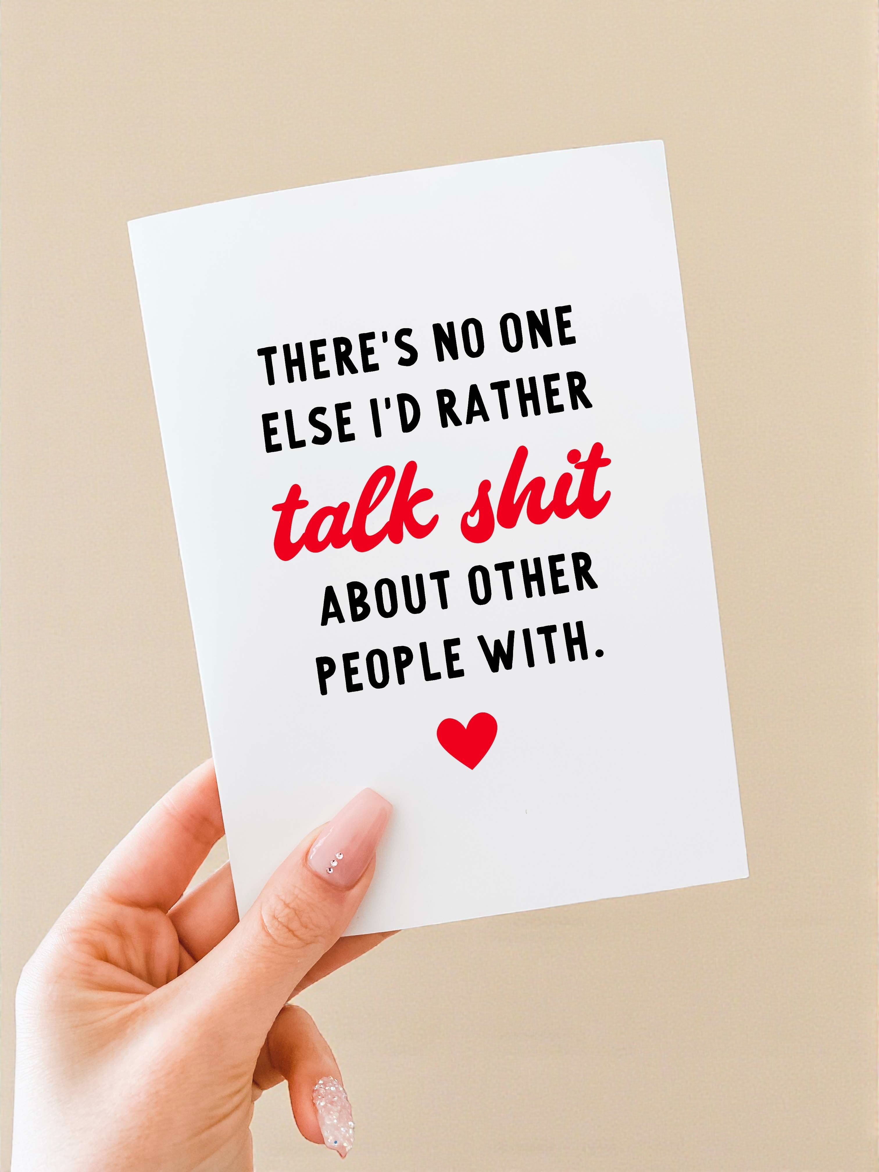 Greeting card held in a hand against a neutral background. The card has a white background with bold black text that reads, “There’s no one else I’d rather,” followed by the phrase “talk shit” in red script-style lettering, and then black text reading, “about other people with.” A small red heart icon appears centered near the bottom of the card. The card is vertically oriented and blank inside