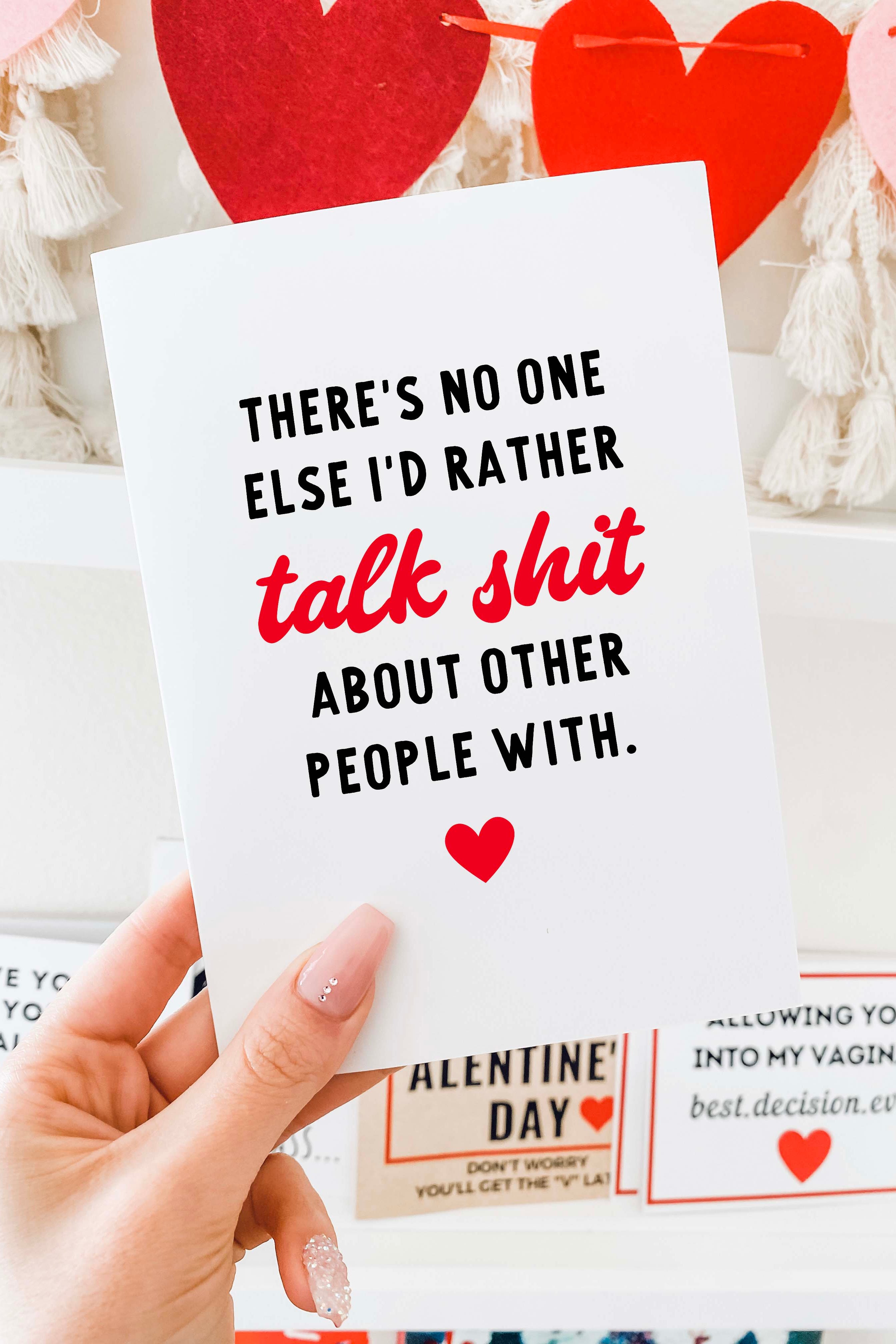 A greeting card is held in a hand against a shelf display with red and pink heart decorations in the background. The card has a white background with black text that reads, “There’s no one else I’d rather,” followed by the phrase “talk shit” in red script lettering, and then black text reading, “about other people with.” A small red heart icon is centered near the bottom of the card. The card is vertically oriented and blank inside.