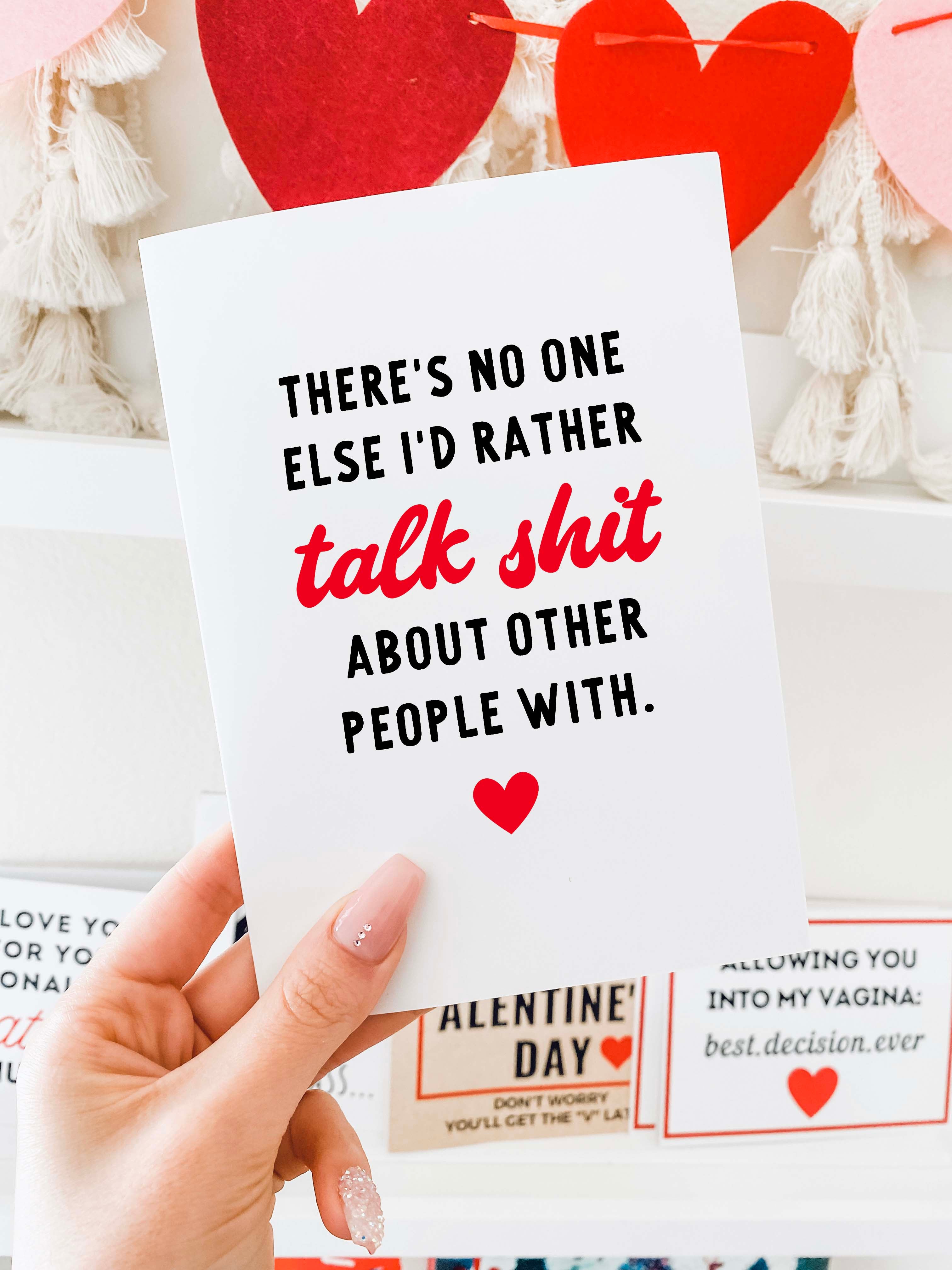 A greeting card is held in a hand against a shelf display with red and pink heart decorations in the background. The card has a white background with black text that reads, “There’s no one else I’d rather,” followed by the phrase “talk shit” in red script lettering, and then black text reading, “about other people with.” A small red heart icon is centered near the bottom of the card. The card is vertically oriented and blank inside.
