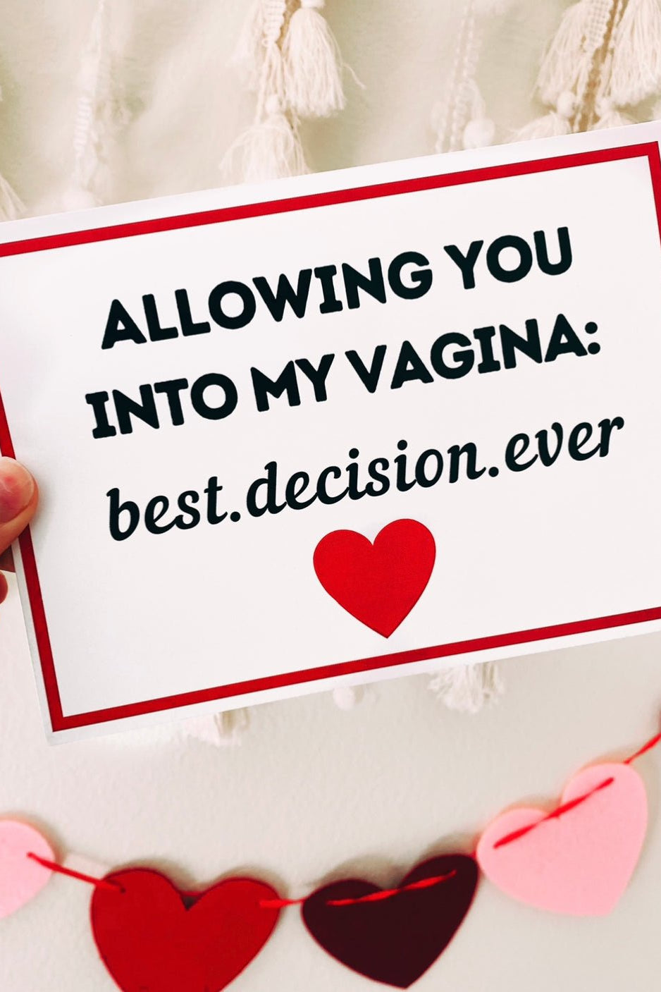 A hand holding a white greeting card with a red border and black text that reads “Allowing you into my vagina: best decision ever,” with a red heart printed below the text. The background includes hanging tassels and a garland of red and pink heart shapes.
